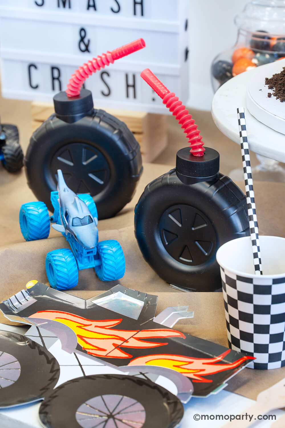 A Monster Truck bash party Table styled by Momo Party featuring a black monster truck shaped paper plates, a checker board party cup and matching checkered paper straw, with big tire shaped tumbler cups with red plastic straws, on a brown kraft table runner decorated with monster truck toys. In the back there's a sign says "Smash & Crush" a fun table setting for kid's monster truck themed birthday party.