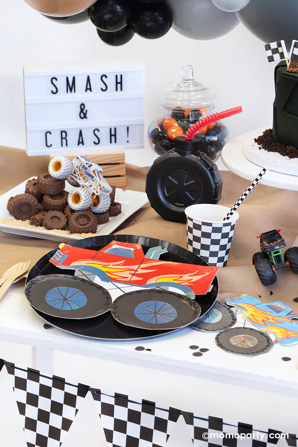 A monster truck themed party table setting styled by Momo Party decorated with party balloon cloud and checkerboard race car flags banner. The table features a red monster truck shaped paper plate with black round dinner plate, a truck shaped blue napkin, a checkerboard party cup and matching paper straw, a tire shaped tumbler cup with red straw on a brown craft table cover decorated with monster truck toys and black confetti. In the back there's a "smash & crush" sign, a fun setup for kid's birthday party.