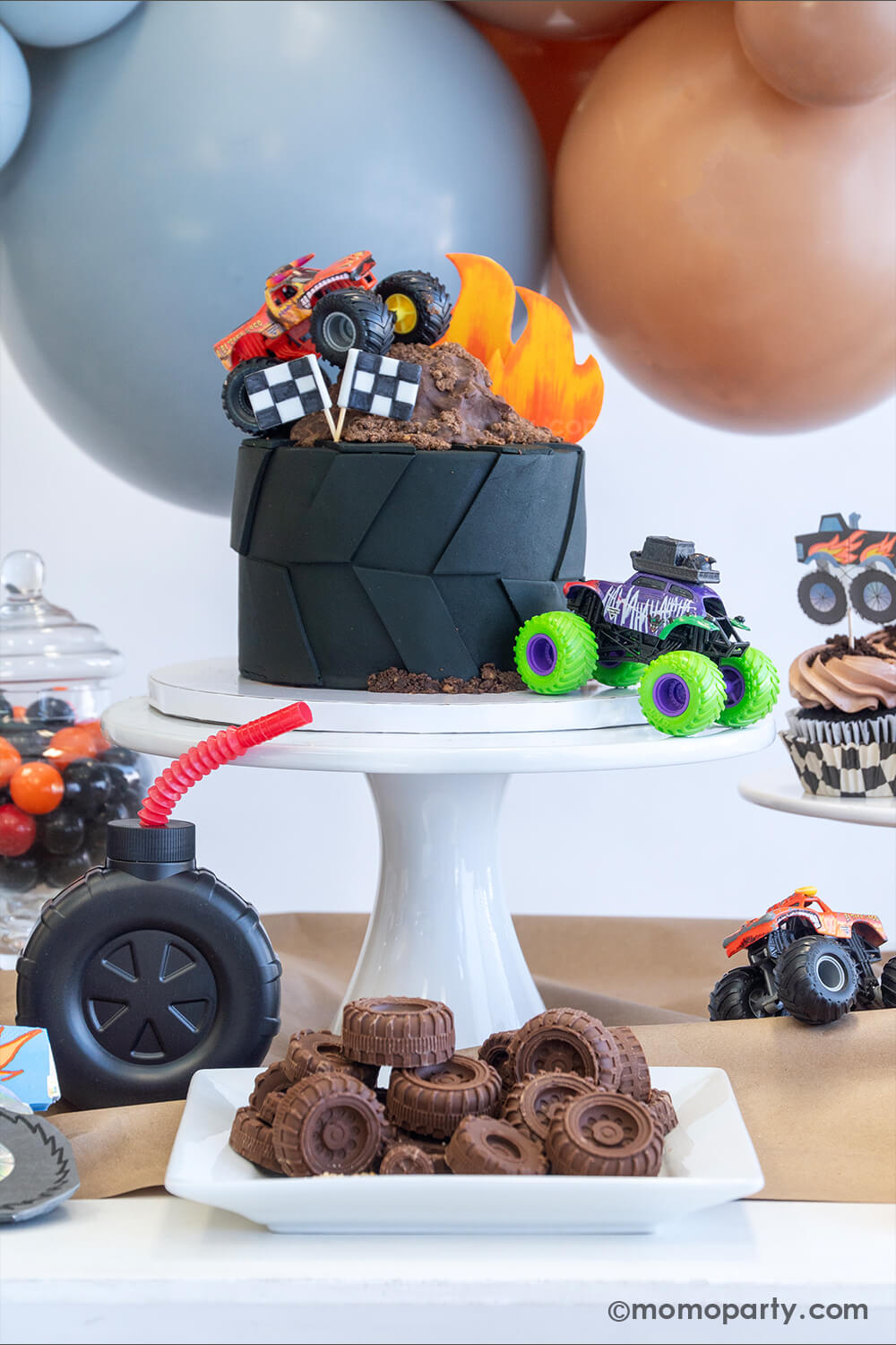 A monster truck themed party table setting styled by Momo Party. The table features a large fondant tire shaped birthday cake topped with a fondant flame, chocolate mud, two race car flags and two colorful monster truck toys. In the front there's a plate full off tire shaped chocolate candies inspired by monster truck tires. The table is covered with brown kraft paper with a candy jar with black and orange gum balls, a tire shaped tumbler cup with a red straw and a few colorful monster truck toys on it, fun