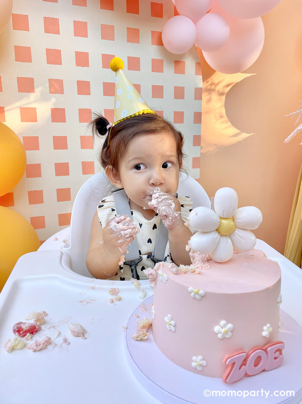 Momo Party One Groovy Baby First Birthday Party, with a birthday girl wearing a Retro Daisy Party Hat, sitting in the hair chair, eating her modern groovy-themed light pink smash cake decorated with Dasiy flower topper and Zoe in the front. She sitting in front of a check-patterned wooden Backdrop board decorated with latex balloons in peach, pink, and Goldenrod colors. This decoration is such a stand-out backdrop and photo booth for a Groovy birthday party, baby showers, and almost any other joyful event