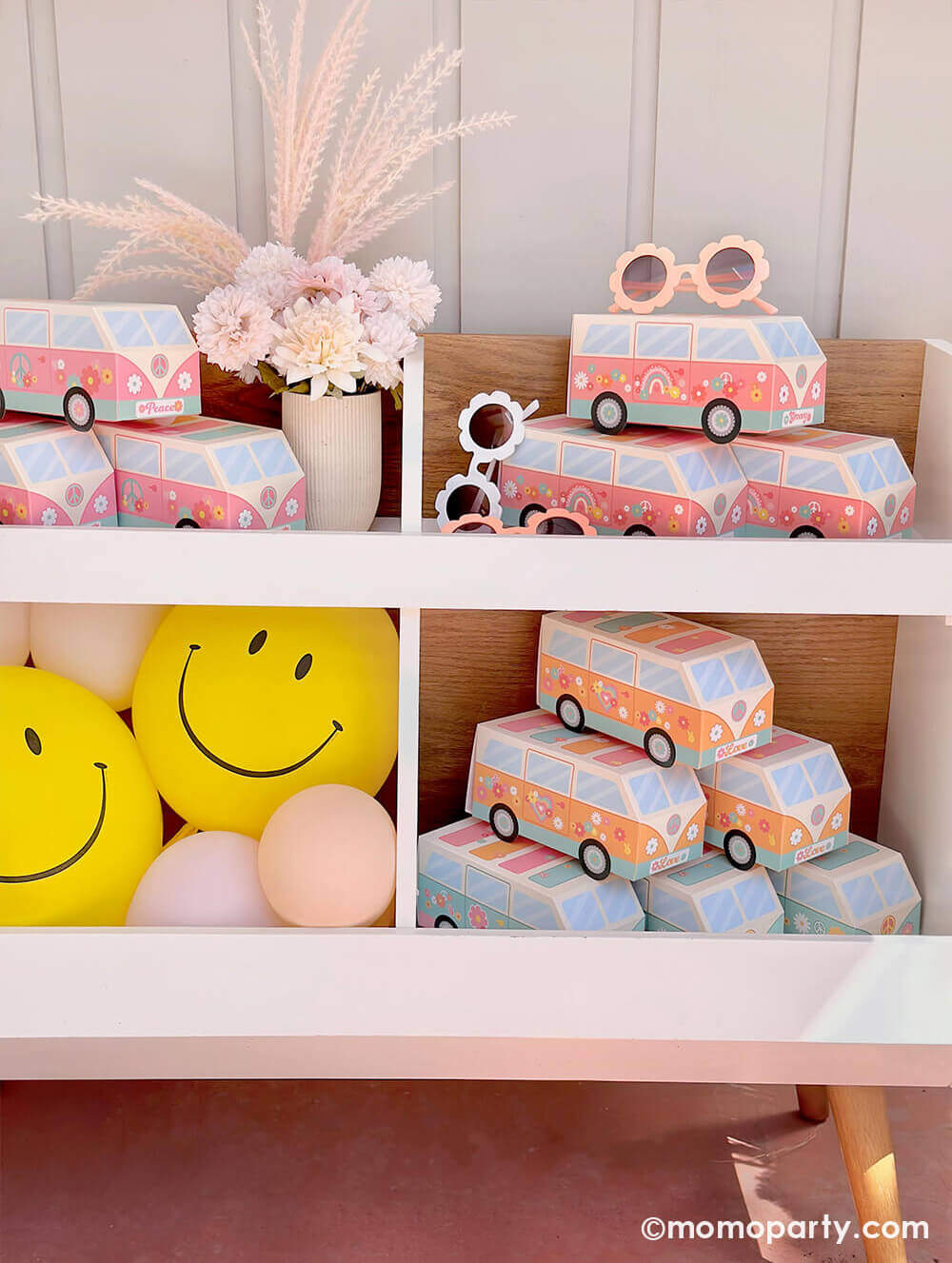 A book shelf filled with hippie themed VW van shaped treat boxes in pink and orange, along with Momo Party's yellow smiley face latex balloons at a baby girl's "Groovy One" themed first birthday party.