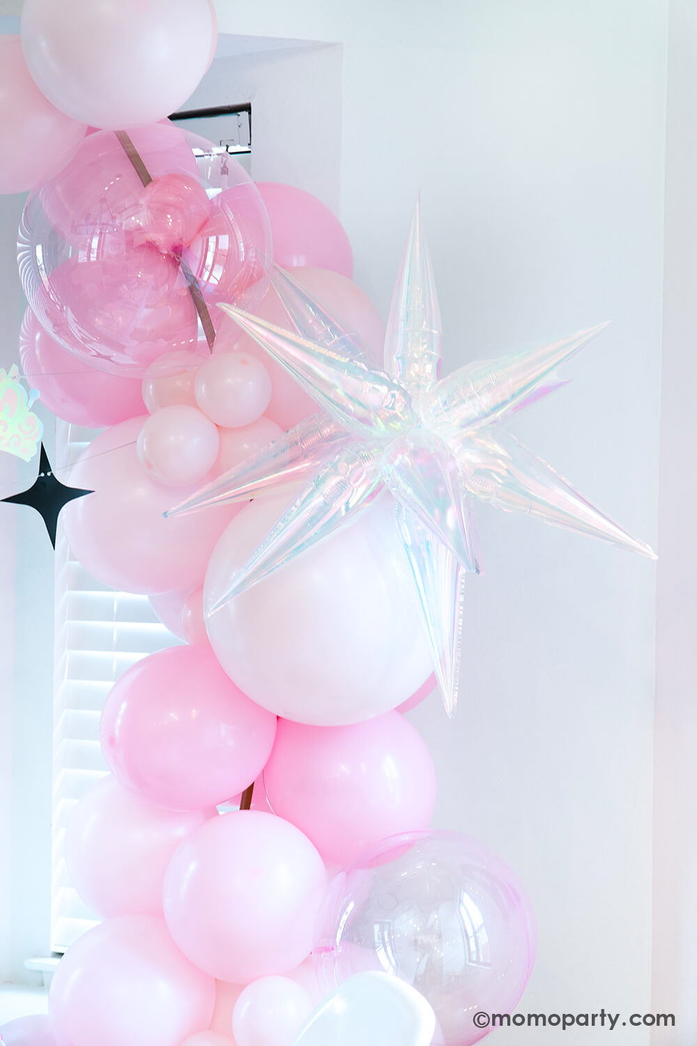 A Wicked Glinda inspired balloon arch by Momo Party with pink and pastel pink latex balloons in various sizes adorned with the 26" iridescent starburst shaped foil balloon and two clear pink orbz non-foil balloons as bubbles next to a window against a white background at a Wicked themed party.