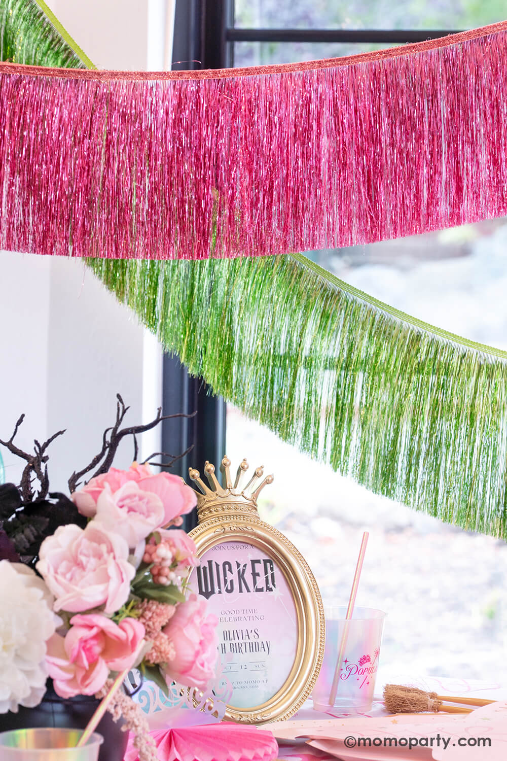 A Wicked themed party decoration set up featuring the pink and green tinsel fringe garlands from Momo Party hung above the party table with 'Wicked' themed photo frame and flowers arrangement.