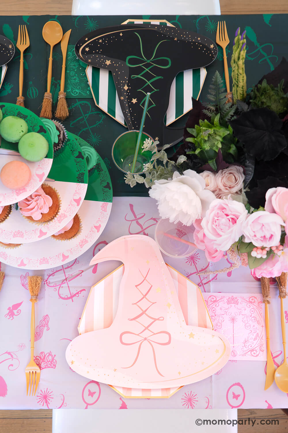 A Wicked themed party table setting with various of Momo Party's Wicked themed party supplies including witch hat shaped plates in pink and black, napkins inspired by Glinda and Elphaba, witch broom shaped utensils, and Wicked Emerald City 3 tier cupcake stand on the pink and green two-tone Wicked tablecloth. With Wicked inspired floral bouquet, it makes a whimsical and mystical Halloween celebration in this spooky season!