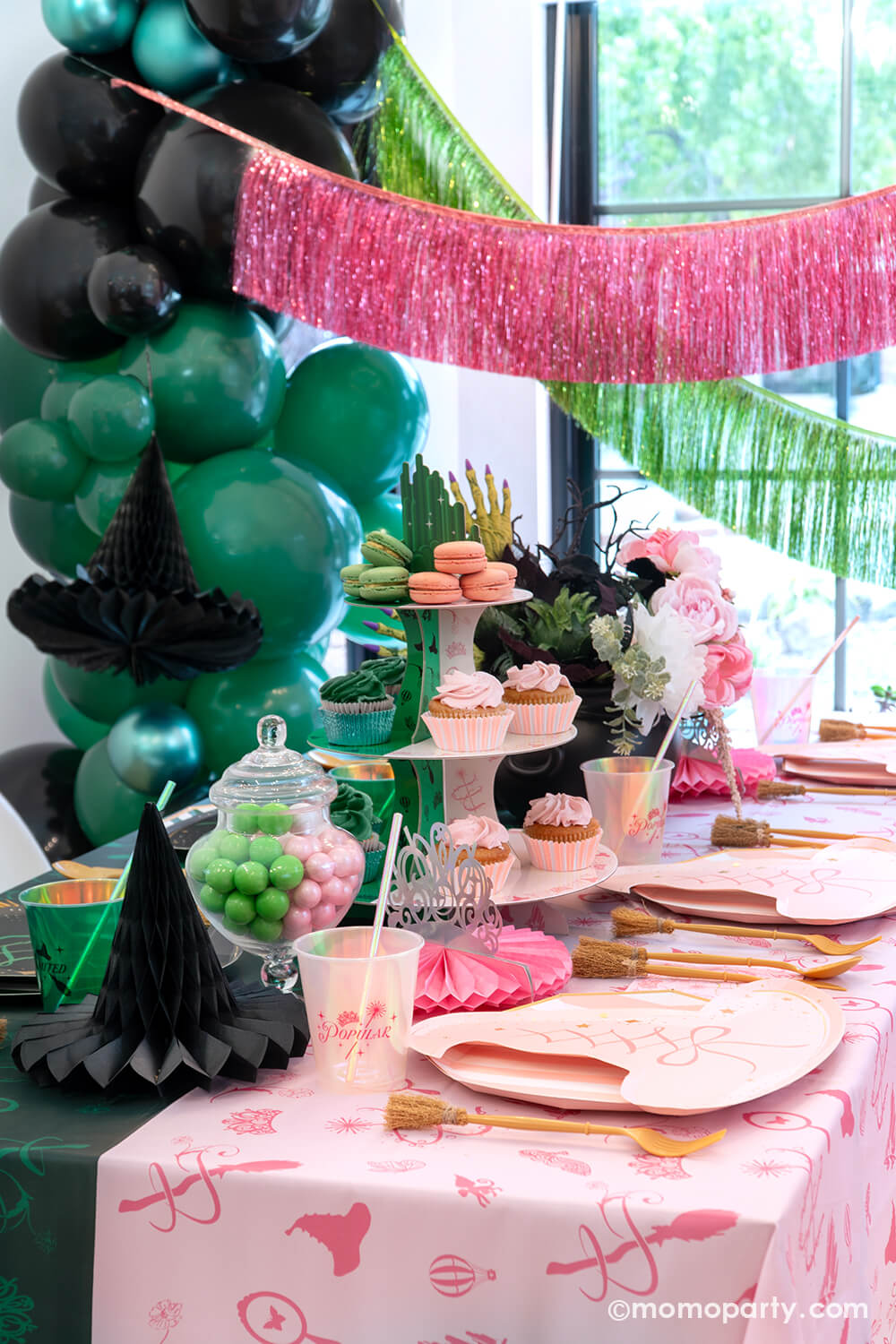 A Wicked inspired party table setting featuring various Wicked themed party supplies and decorations from Momo Party including pink and black witch hat shaped plates, pink and green two-toned Wicked Emerald City cupcake stand, Wicked party cups, witch broom utensil set and Wicked witch hat honeycombs on the green and pink two-tone tablecloth. With green and pink floral bouquet and treats, balloons and fringe garlands in the backtground, this makes a mystical yet whimsical table for a Wicked party!