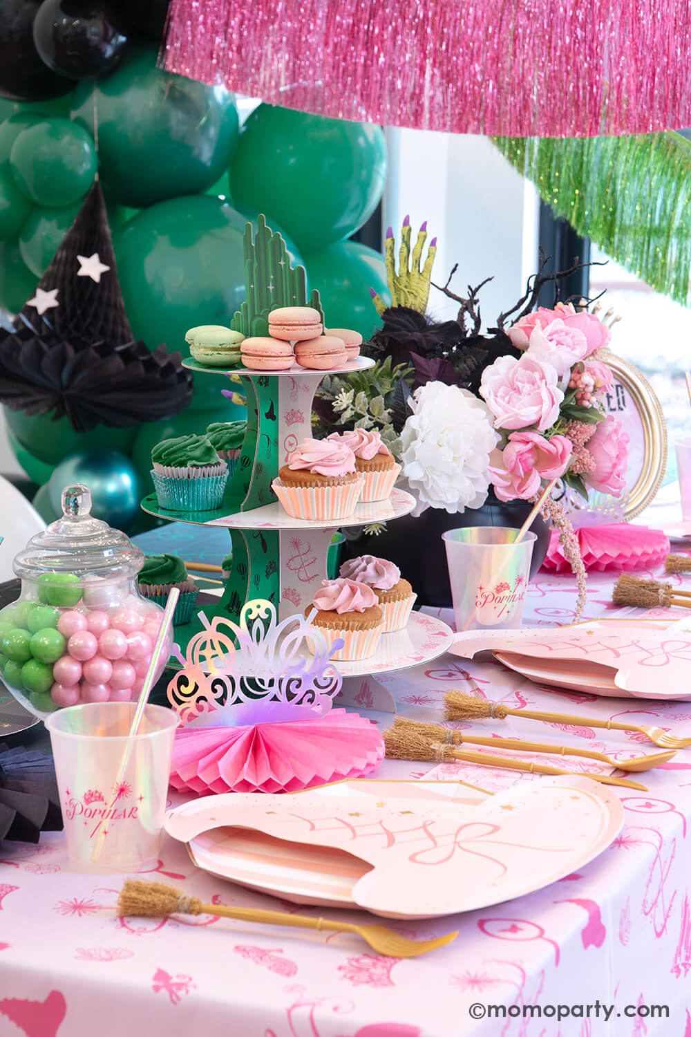 A Glinda inspired Wicked themed party table setting featuring various Wicked themed party supplies and decorations from Momo Party including Glinda pink tiaras, pink witch hat shaped plates, pink and green two-toned Wicked Emerald City cupcake stand, Glinda iridescent party cups, witch broom utensil set and Wicked witch hat honeycombs on the pink tablecloth. With green and pink floral bouquet and treats, this makes a mystical yet whimsical table for a Wicked themed party this spellbinding season!