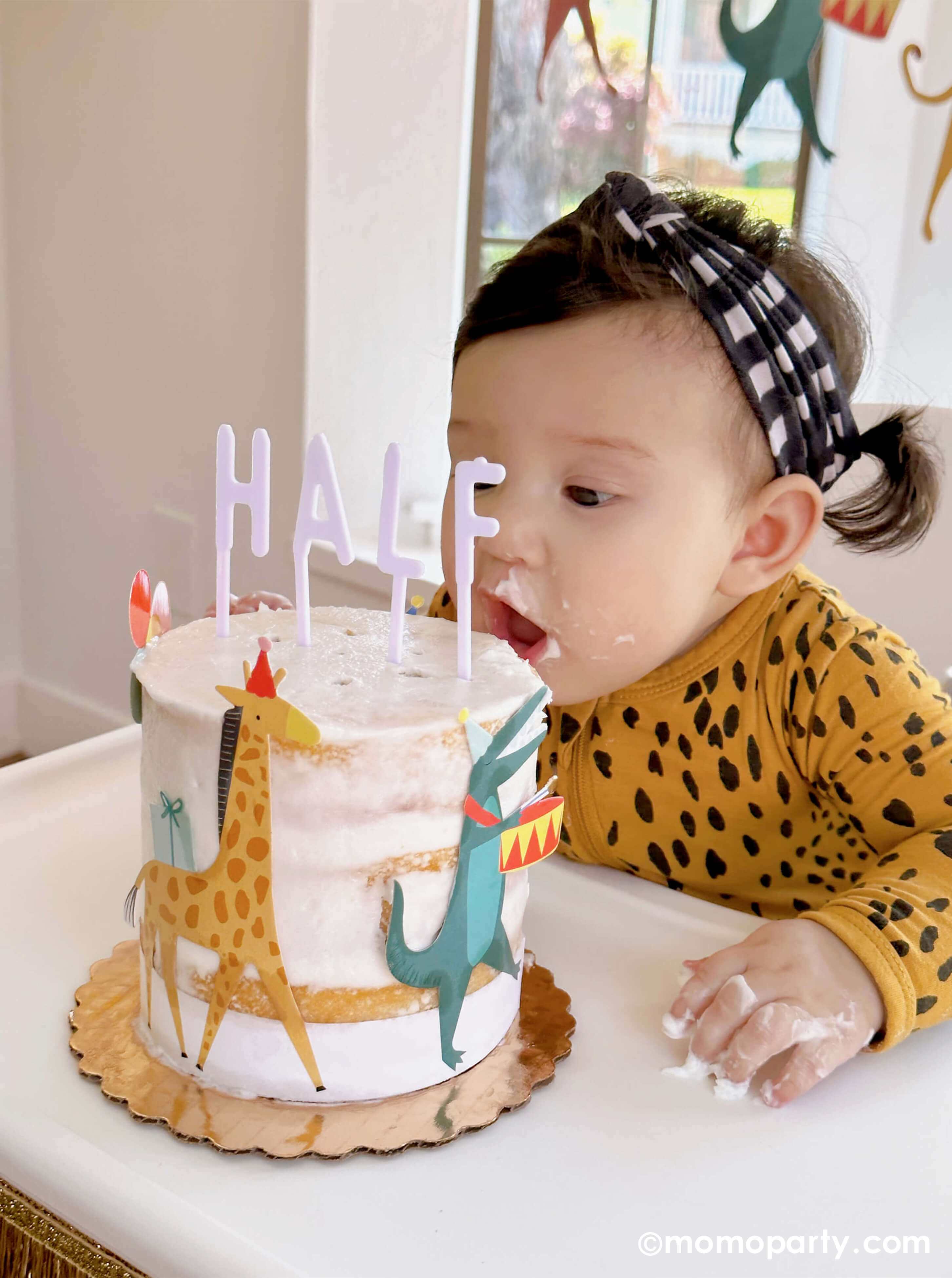 A cute baby girl wearing animal pattern onesie, sitting on a highchair in her 6 months animal themed half birthday party, garbing her Naked Cake decorated with Meri Meri Animal Parade Cake Wrap & Toppers, and on the top spelling "HALF" with Cake By Courtney Letter-board Cake Toppers, repersend for a six months half birthday, along with Meri Meri gold fringe garland around the highchair tray, it makes a great party inspiration for kid's animal, safari, carnival themed birthday party.