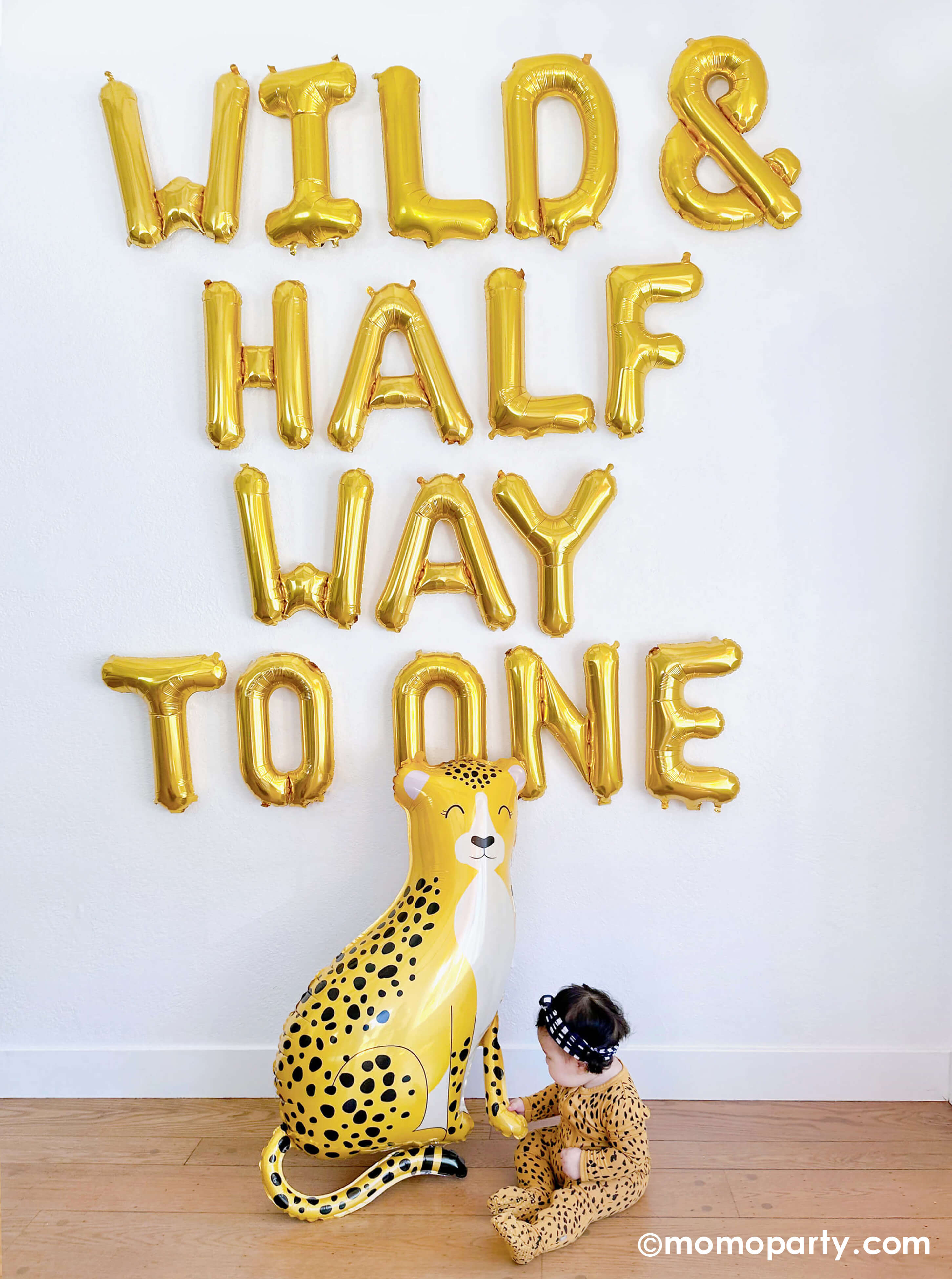 A balloon wall featuring Momo Party's "Wild & Half Way To One" 16" Mylar Letter Balloons by Northstar Letter Balloons. At the bottom of the wall sits Momo Party's baby cheetah shaped foil balloon by Betallic Balloons and a 6-month baby girl in her cheetah print onese reaching out her hand to hold the cheetah, all makes a great inspiration for an adorable baby's half birthday party.