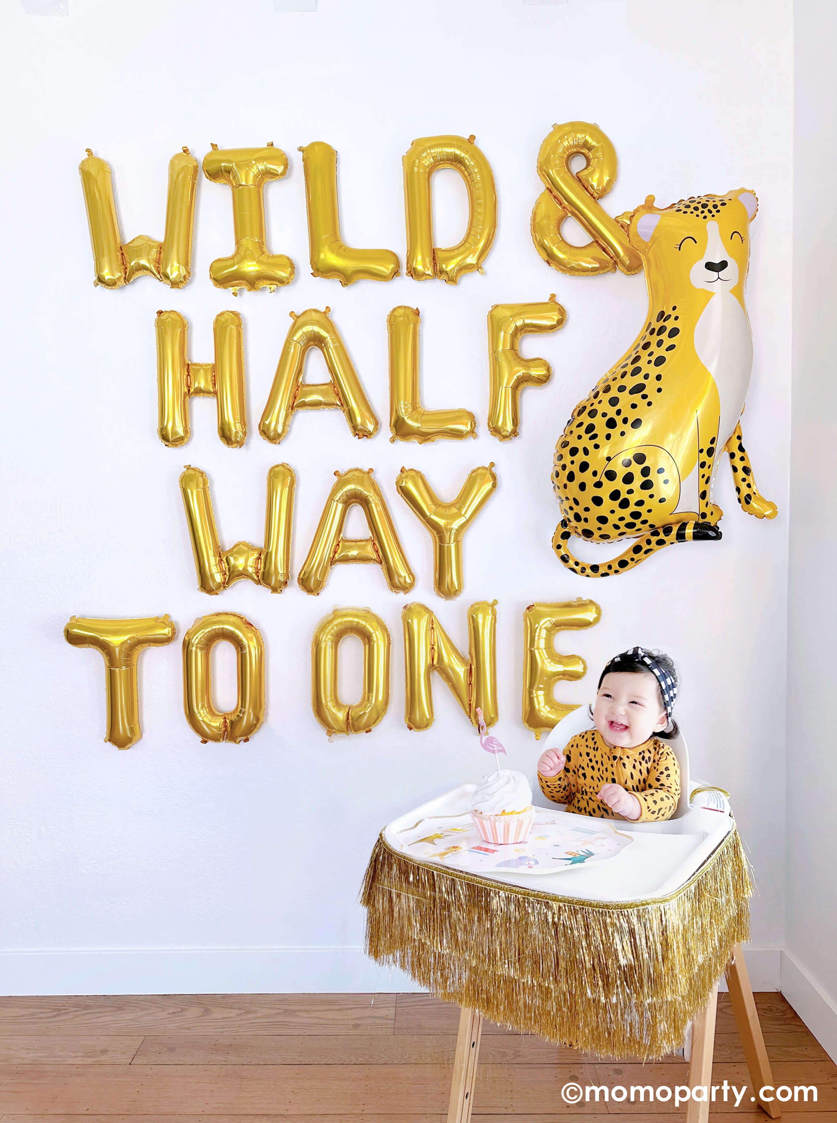 A balloon wall featuring Momo Party's Wild & Half Way To One" 16" Mylar Letter Balloons by Northstar Letter Balloons and Momo Party's 40" baby cheetah shaped foil balloon by Betallic Balloons. Next to the balloon wall, a happy smiley 6-month baby girl in her cheetah print onese sits in a highchair decorated with Meri Meri's gold tinsel garland, on her tray, a cupcake topped with a cute flamingo shaped topper , all makes a great inspiration for an adorable baby's half birthday party.