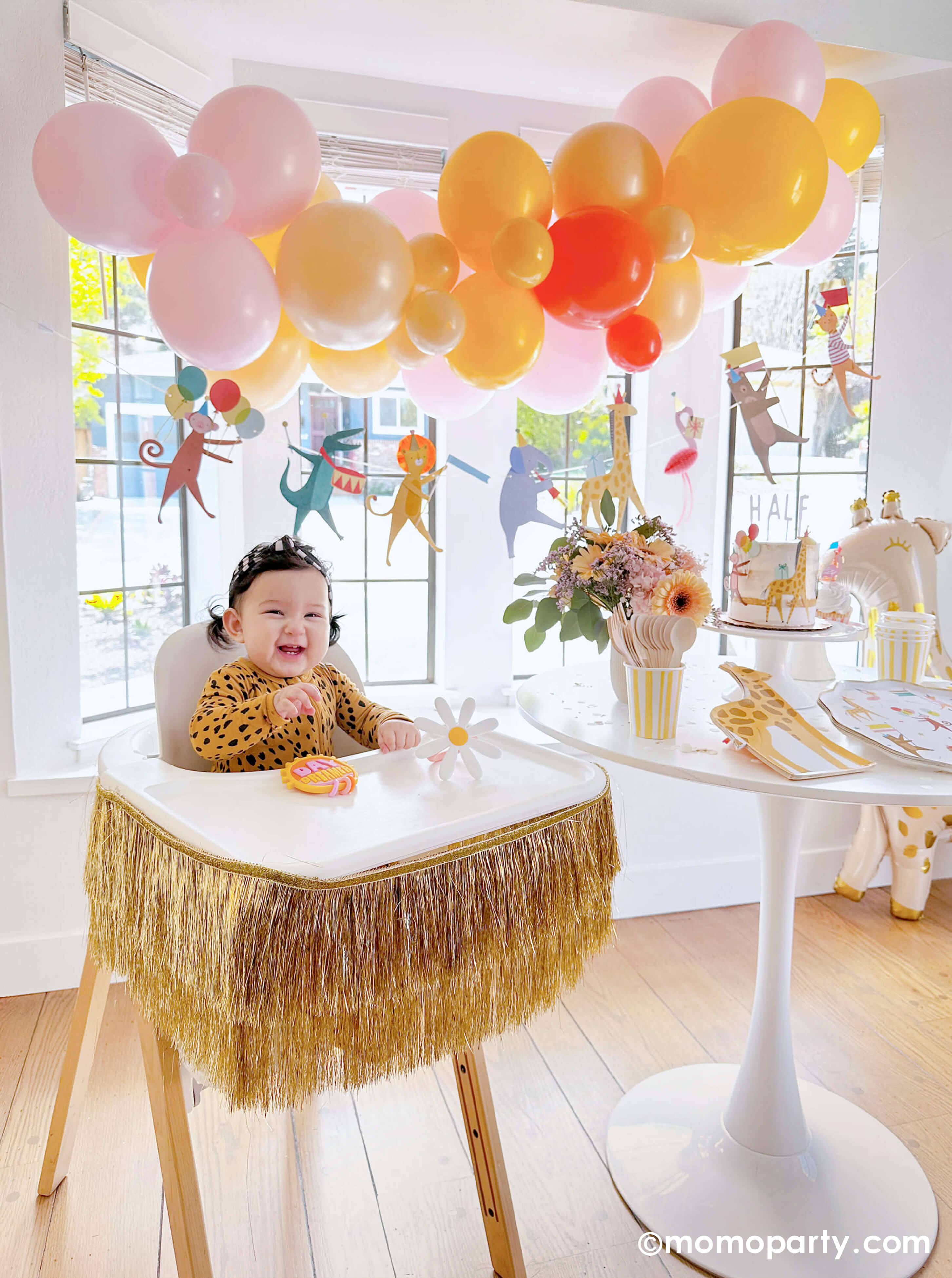 A happy baby girl's 6 months animal themed birthday party featuring Momo Party's animal parade birthday garland featuring a lion, an elephant, an alligator, a giraffe, along with a festive balloon garland in a warm color tone hanging above. On the dessert table, there are Meri Meri's animal parade dinner plates, giraffe shaped plates and cupcakes dressed with the safari animal toppers, next to the table there's a giraffe shaped foil balloon. One the left a highchair is decorated with a gold tinsel garland.