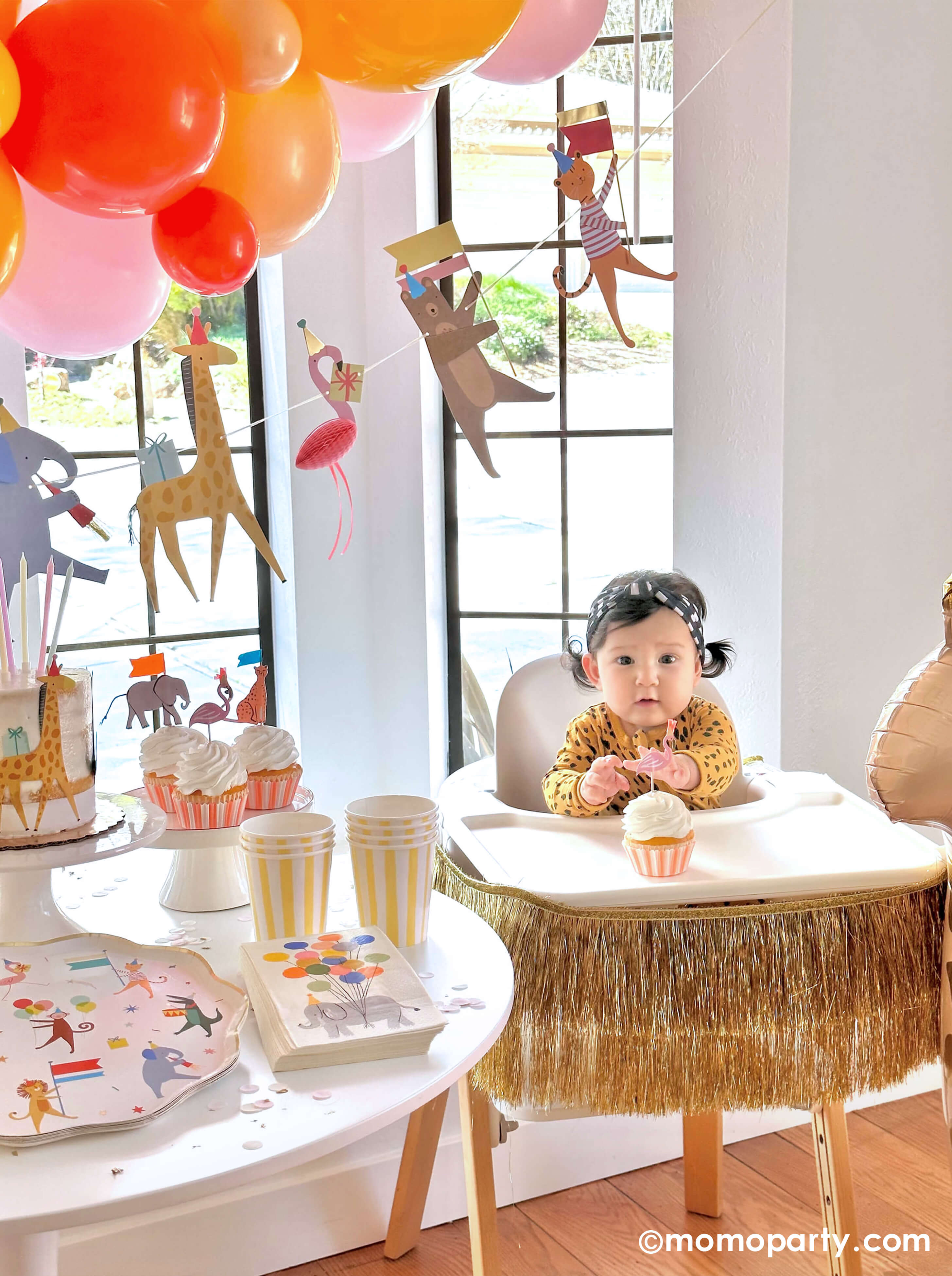 A cute baby girl wearing animal pattern onesie, sitting on a highchair in her 6 months animal themed half birthday party, reaching a cupcake decorated with Meri Meri Safari Animals Cupcake Kit, next to a party table featuring Momo Party's Animal Parade Dinner plate, Rifle Paper Co's party animal guest napkins, a cake decorated with  , along with light yellow striped cups and yellow wooden utensil.  it makes a great party inspiration for kid's animal, safari, carnival themed birthday party.