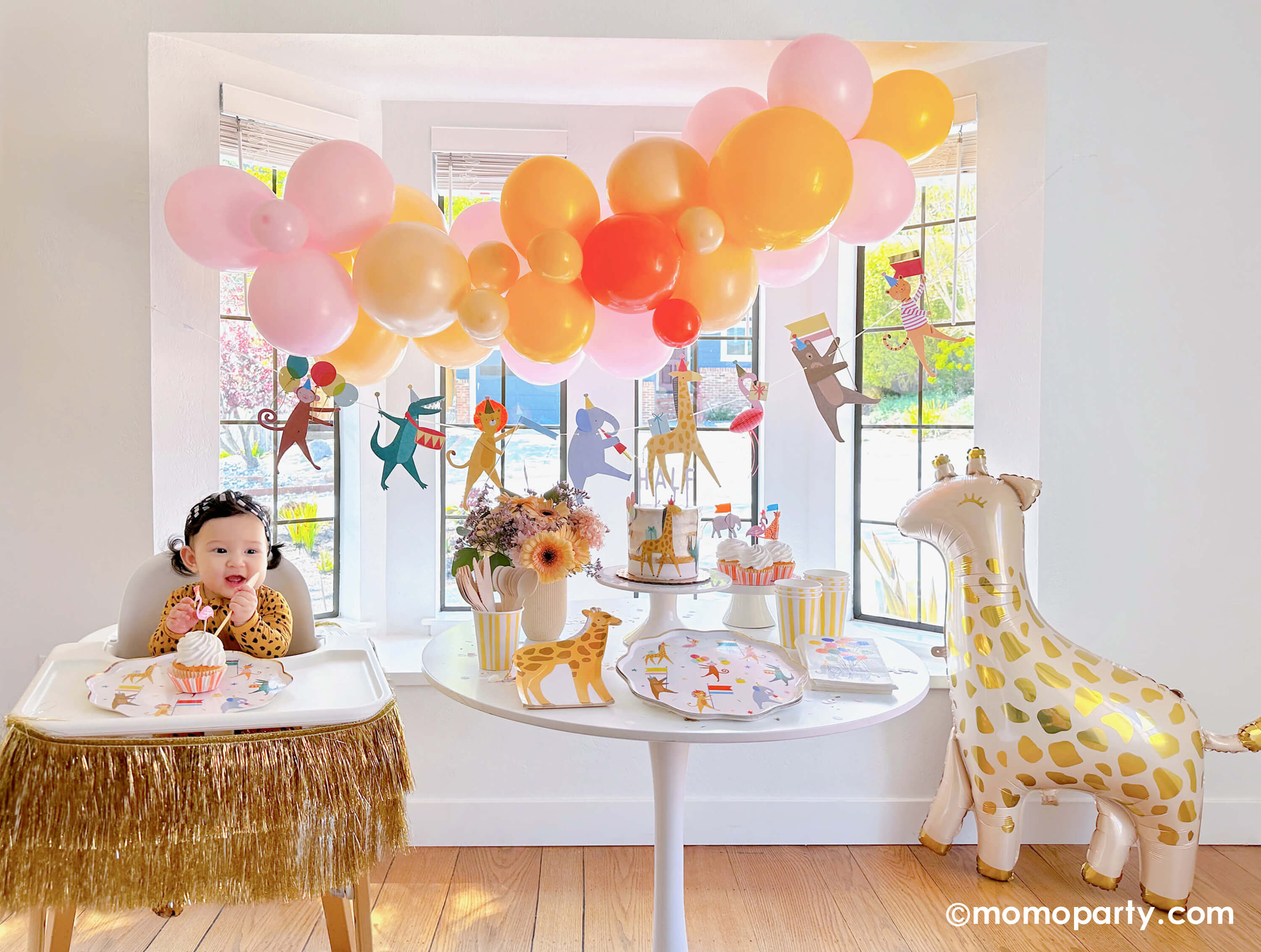 An adorable baby's animal themed 6 months party featuring Momo Party's animal parade birthday garland featuring a lion, an elephant, an alligator, a giraffe, along with a festive balloon garland in a warm color tone hanging above. On the dessert table, there are Meri Meri's animal parade dinner plates, giraffe shaped plates and cupcakes dressed with the safari animal toppers, next to the table there's a giraffe shaped foil balloon. One the left a highchair is decorated with a gold tinsel garland.