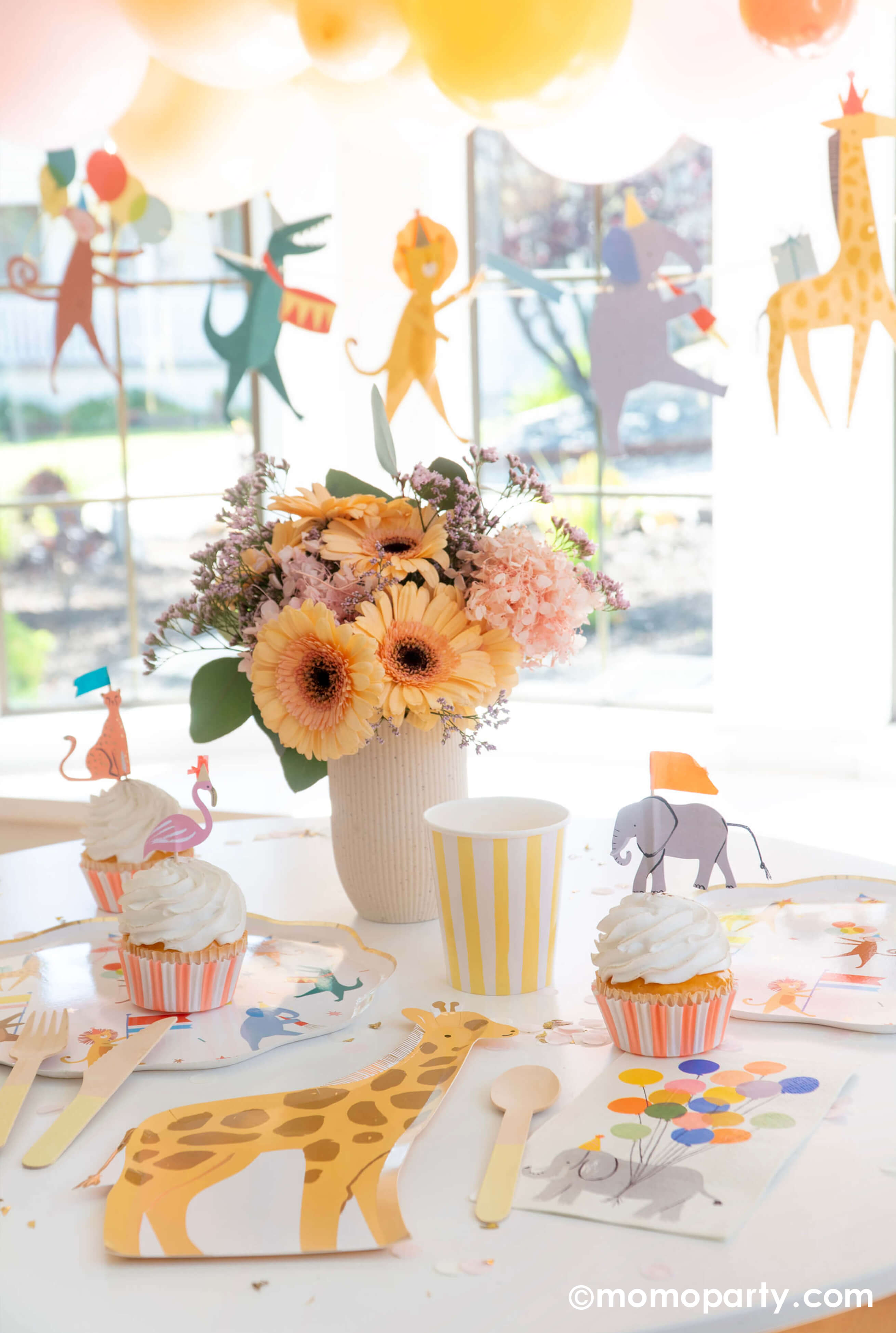 A kid's animal themed party table featuring Momo Party's Animal Parade Dinner plate, a giraffe shaped plate, cupcakes with adorable safari animal cupcake toppers by Meri Meri, Rifle Paper Co's party animal guest napkins flowers in a white vase, along with light yellow striped cups and yellow wooden utensil. There is also a Animal Parade Garland with balloons decorated behind the table. it makes a great party inspiration for kid's animal, safari, carnival themed birthday party.