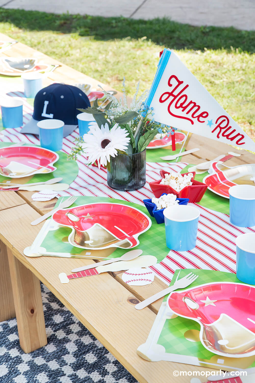 A kid's baseball themed birthday party picnic table setting styled by Momo Party featuring various baseball themed tablewares from Momo Party including baseball helmet shaped plate, cornflower tumbler cups, baseball shaped napkins on the baseball diamond placemats with the red pinstriped runner underneath. In the center there's a beautiful flower bouquet with the "Home Run" felt party pennant and some star shaped trays in blue and red filled with popcorn.