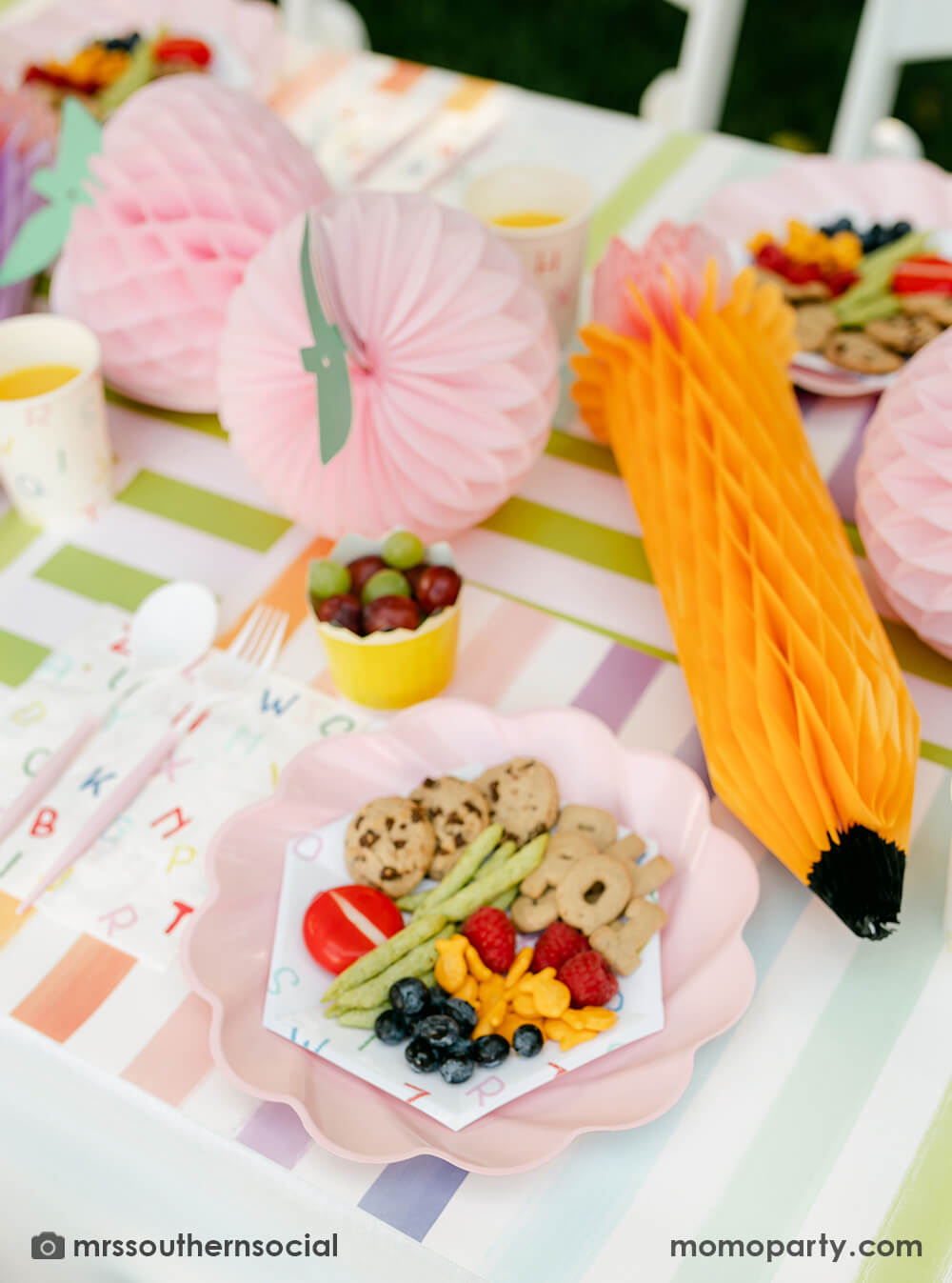 A pastel back to school party table featuring Momo Party's back to school party supplies including ABC alphabet plates, napkins party cups, and pencil shaped food cups. One the plates are kids snack ideas including fruits, cheeses, goldfish, ABC alphabet crackers. With pastel rainbow striped placemats and pastel rainbow pencil honeycombs and pink apple honeycombs, all together makes an adorable tablescape for kid's first day of school celebration.