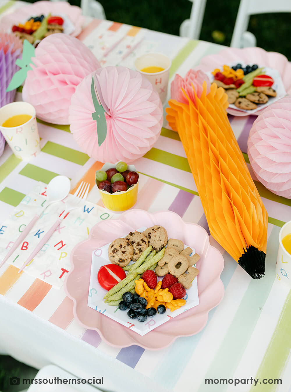 A pastel back to school party table featuring Momo Party's back to school party supplies including ABC alphabet plates, napkins party cups, and pencil shaped food cups. On the plates are kids snack ideas including fruits, cheeses, goldfish, ABC alphabet crackers. With pastel rainbow striped placemats and pastel rainbow pencil honeycombs and pink apple honeycombs, all together makes an adorable tablescape for kid's first day of school celebration.