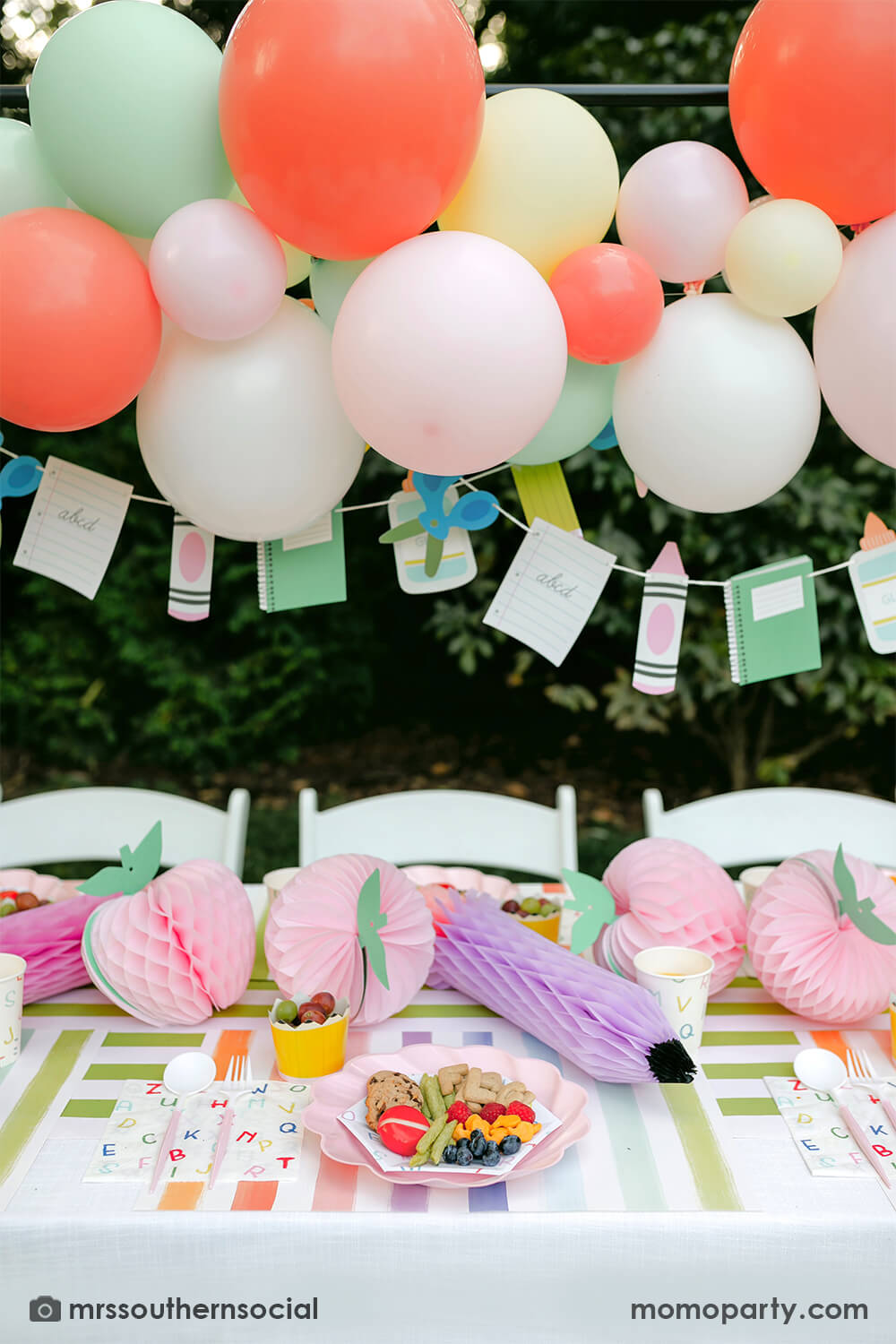 A backyard pastel themed back to school party table set up and decoration featuring Momo Party's back to school themed party goods including back to school party garland, pink apple honeycombs and rainbow pastel pencil honeycombs as the centerpiece, ABC alphabet plates, party cups and napkins. With a pastel themed balloon garland hung above the table, it makes an adorable celebration for kid's first day of school.