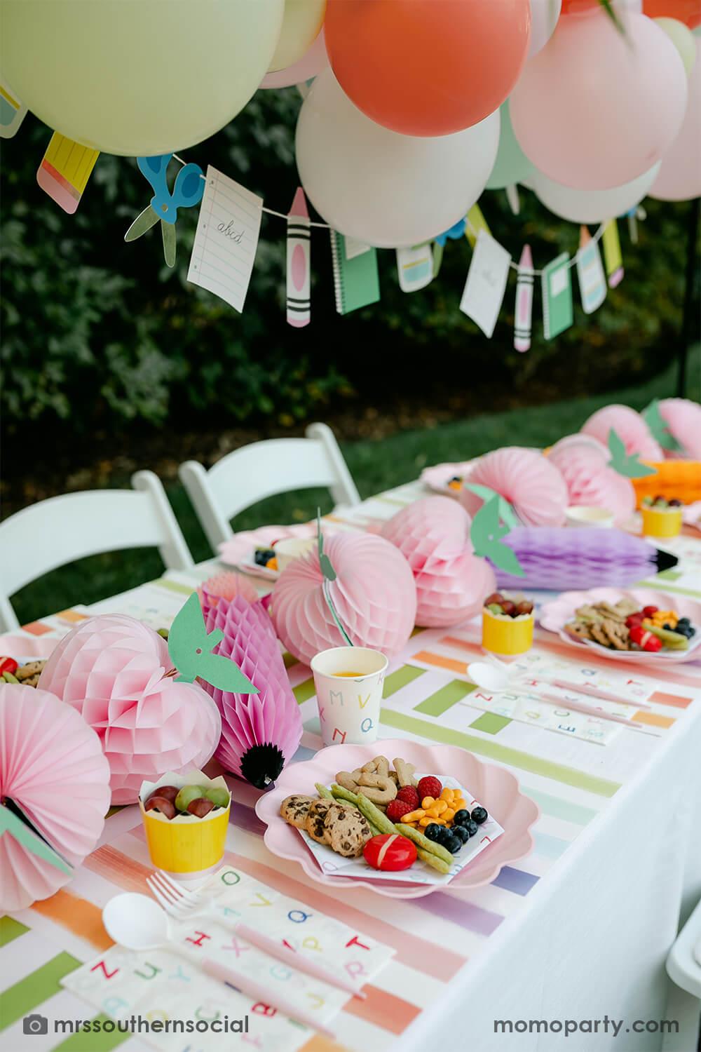 A backyard pastel themed back to school party table set up and decoration featuring Momo Party's back to school themed party goods including back to school party garland, pink apple honeycombs and rainbow pastel pencil honeycombs as the centerpiece, ABC alphabet plates, party cups and napkins. With kids snacks including ABC alphabet crackers, fruits, cheese, it gives great ideas and inspo for celebrating kid's first day of school.
