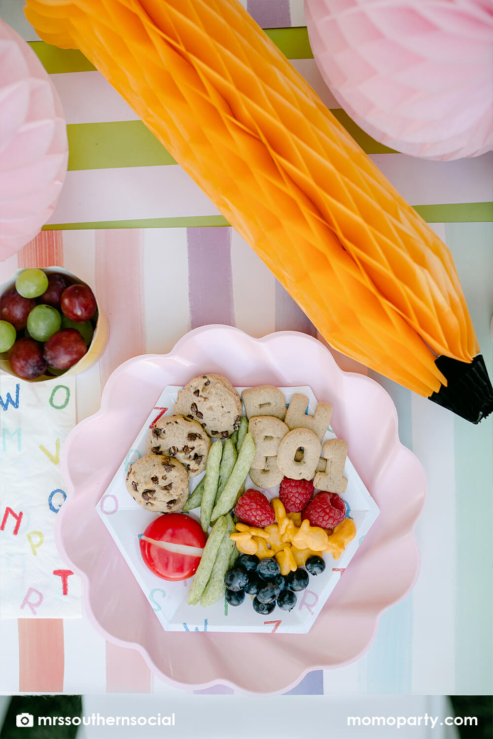 A pastel back to school party table featuring Momo Party's back to school party supplies including ABC alphabet plates, napkins party cups, and pencil shaped food cups. One the plates are kids snack ideas including fruits, cheeses, goldfish, ABC alphabet crackers. With pastel rainbow striped placemats and pastel rainbow pencil honeycombs and pink apple honeycombs, all together makes an adorable tablescape for kid's first day of school celebration.