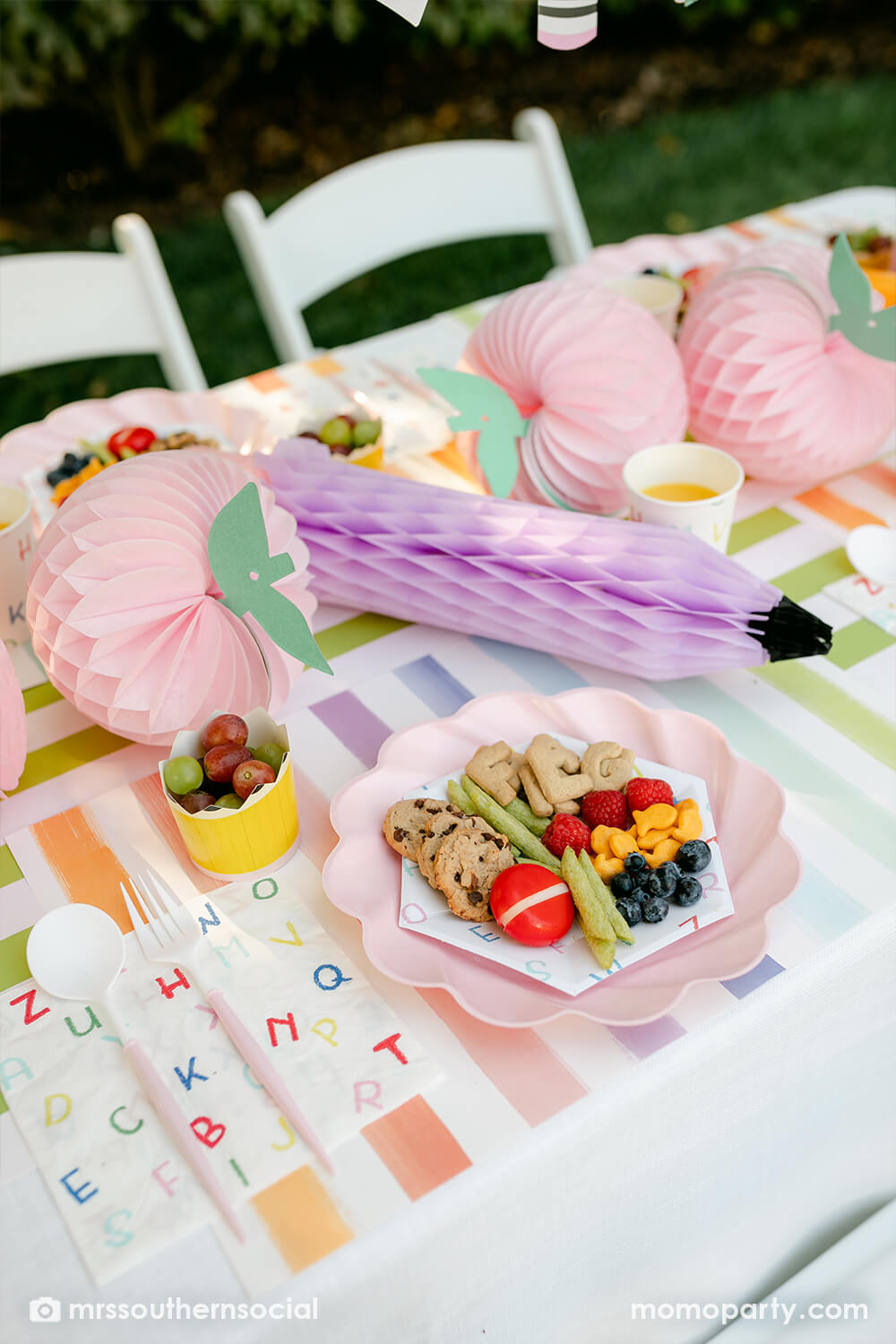 A pastel back to school party table featuring Momo Party's back to school party supplies including ABC alphabet plates, napkins party cups, and pencil shaped food cups. One the plates are kids snack ideas including fruits, cheeses, goldfish, ABC alphabet crackers. With pastel rainbow striped placemats and pastel rainbow pencil honeycombs and pink apple honeycombs, all together makes an adorable tablescape for kid's first day of school celebration.