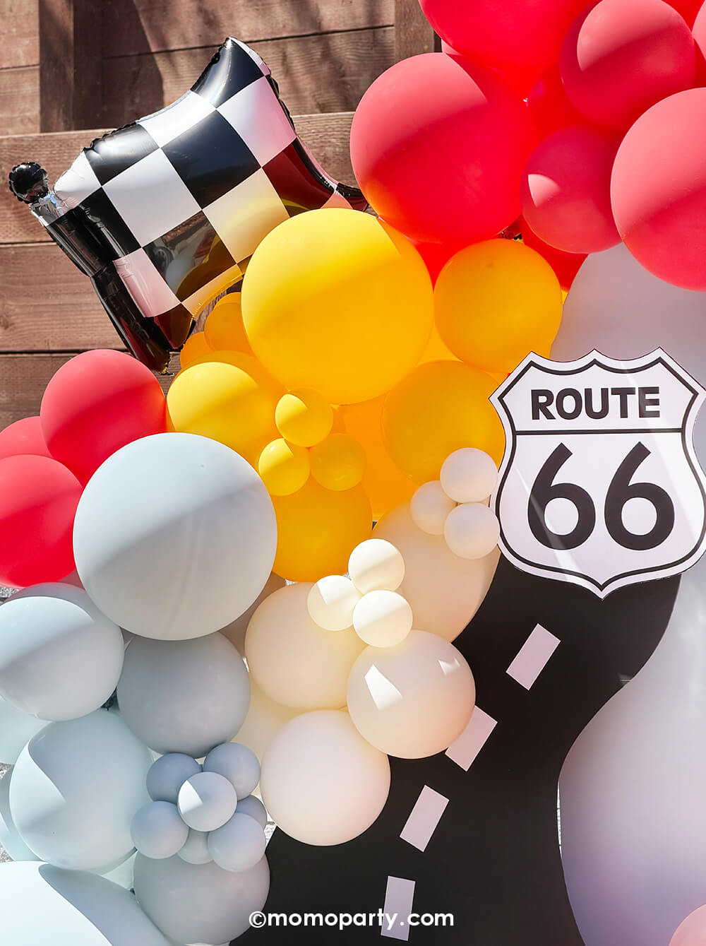 A kid's Disney Cars themed birthday party balloon decoration featuring Momo Party's 16" x 13.8" Checkered Race Car Flag Foil Mylar Balloon, along with a race car themed balloon garland in light blue, red, goldenrod and white colors hung around a party backdrop cutout of a race track and Route 66 cutout sign. A perfect decoration inspo for boy's race car themed birthdays or Lightning Mcqueen themed parties.