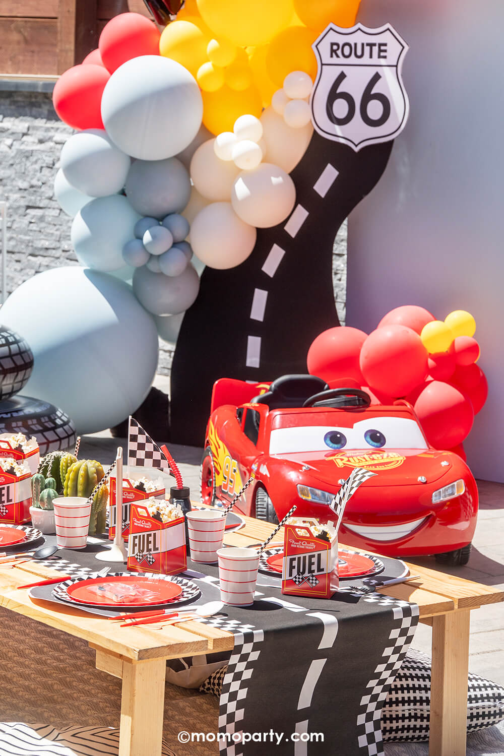 A kid's Disney Cars Lightning Mcqueen themed birthday party set up by Momo Party featuring a table filled with race car themed tableware including race track checkered table runner, fuel treat boxes, red striped cups, checkered plates and mini race car flags as decoration. In teh back is a festive race car themed balloon garland hung around a party backdrop cutout of a race track and Route 66 cutout sign, next to a Lightning Mcqueen race car. A perfect decoration inspo for boy's race car themed birthdays.