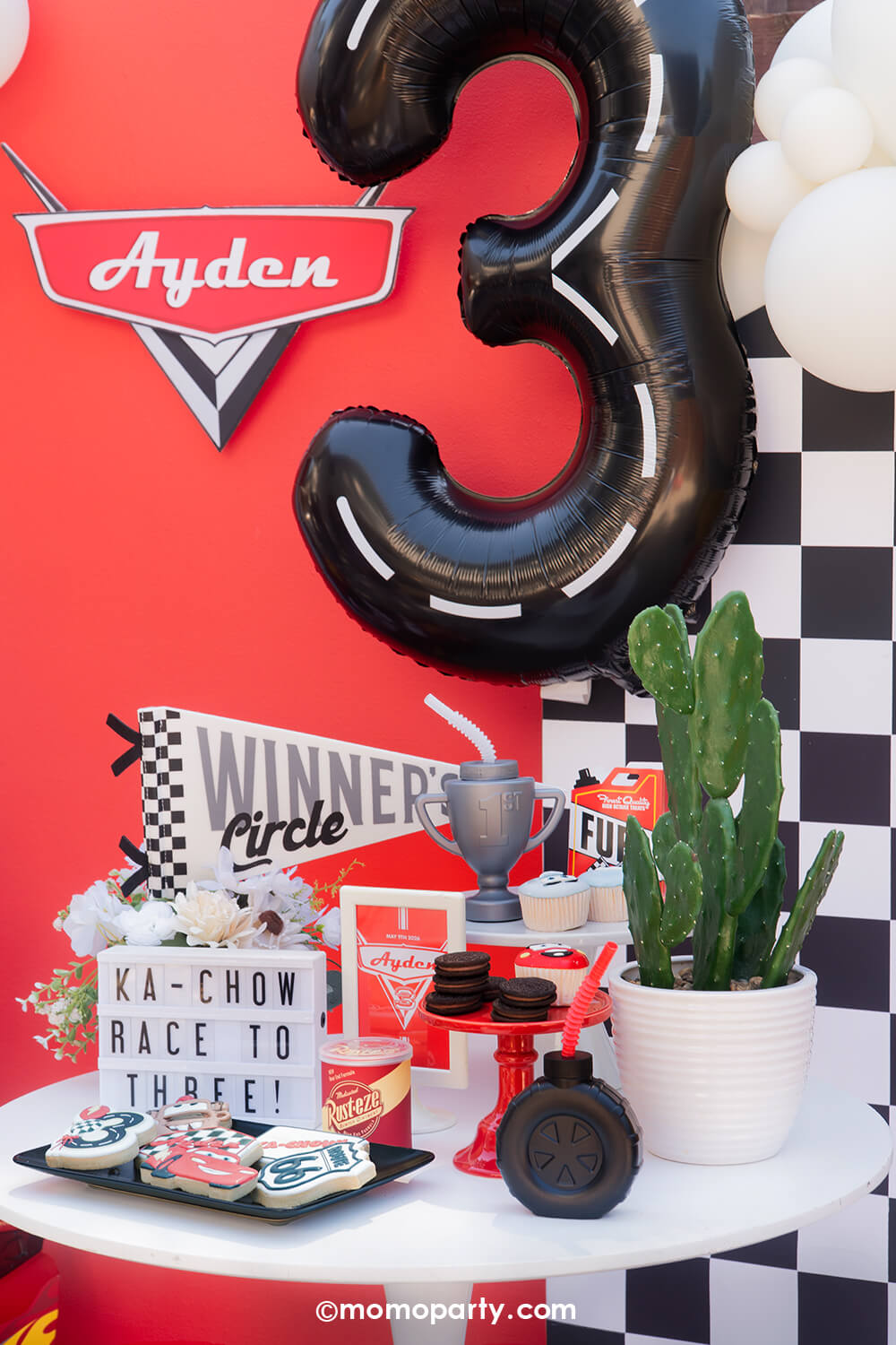 A kid's Disney Cars Lightning Mcqueen themed birthday party table decoration featuring various race car themed party supplies form Momo Party's including the Winner Circle race car felt party pennant, the trophy shaped tumbler, the tire shaped sipper, and fuel gas tin shaped treat boxes. In the background, there's the number 3 road tape foil balloon floating above, along with the custom-made Disney Cars logo and the checkered background, this festive decoration is perfect for boys birthday celebrations!