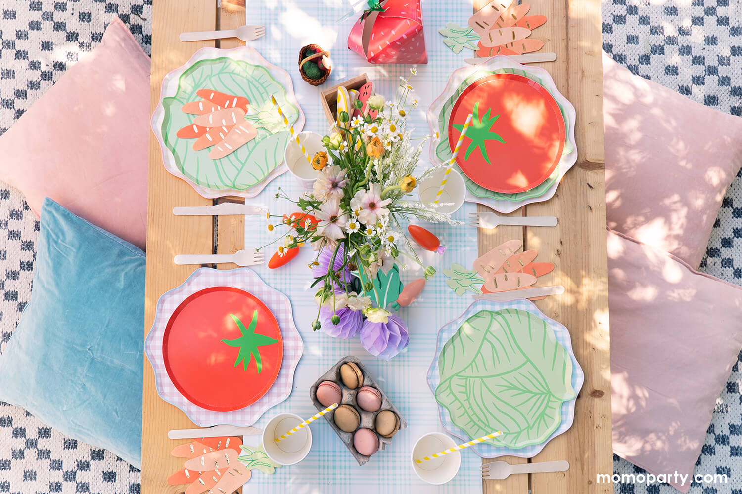 An overhead shot of a farmer's market inspired party picnic table setting by Momo Party featuring tomato shaped paper plates, lettuce shaped plates, carrot bunch shaped napkins and party cups over a light blue gingham runner. In the middle is a floral bouquet along with some sweet treats in mini wooden crate and egg cartons. The picnic table is on a checkered rug adorned with light pink and pastel mint pillows.