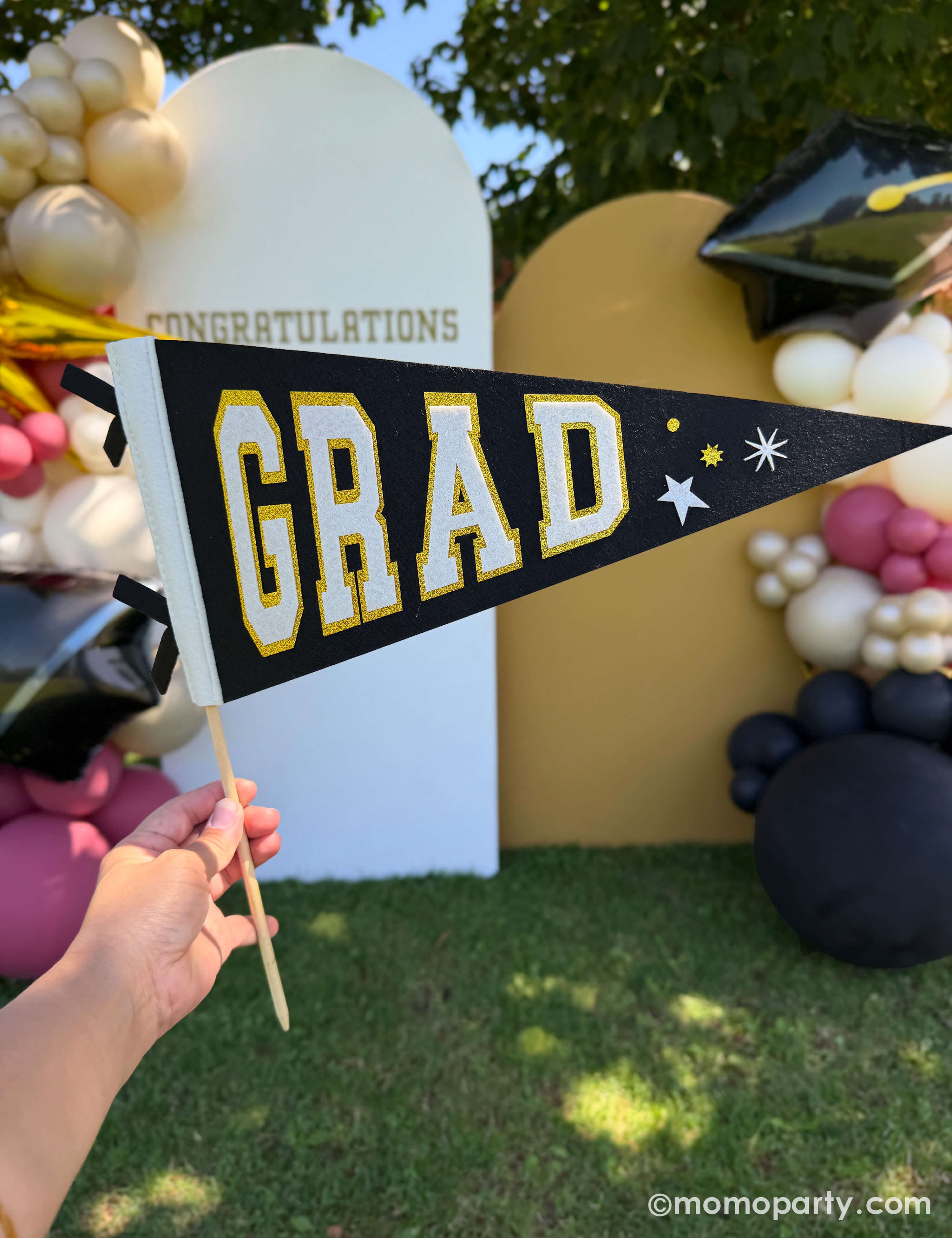 Graduation-themed felt pennant held by a hand in front of a graduation party backdrop adorned with a festive graduation balloon garland with graduate cap shaped foil balloon and 'Congratulations' sign in the background.