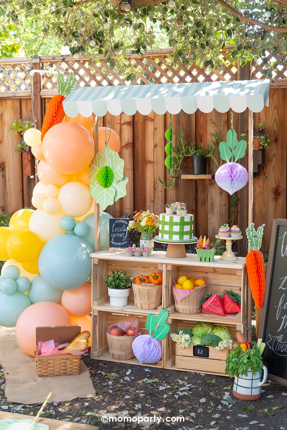 A children's farmer's market themed party outdoor setup by Momo Party with a mini wooden market stall adorned with a colorful pastel balloon garland, produce shaped foil balloons, veggie honeycomb hanging decorations, and food-themed party tablewares on the party table. Along with cakes, cupcakes, sweet treats, floral arrangement, baskets filled with fruits and vegetables, this adorable set up makes a cute kid's birthday party, a baby shower, or a family friendly spring/summer celebration.