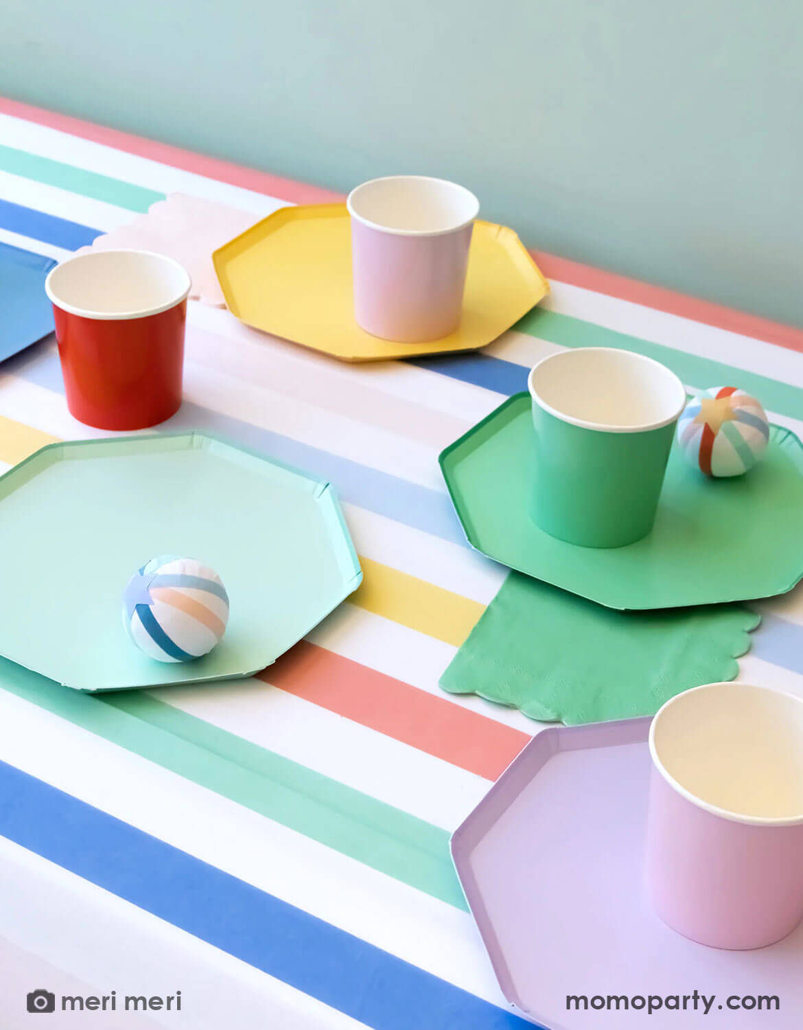 A colorful table setting featuring Meri Meri's multi-color hexagonal party plates, cups, napkins and surprise balls from Momo Party on a multi-color striped tablecloth.
