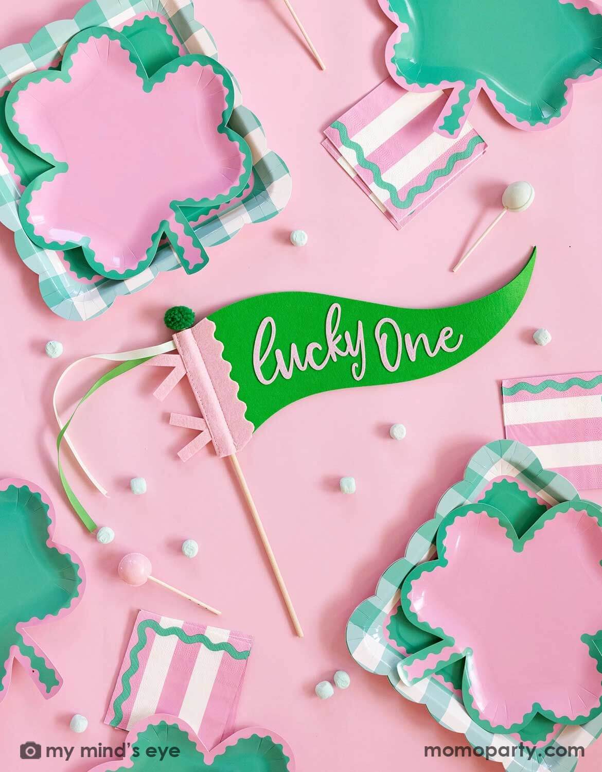 Decorative pink and green party plates with shamrock shapes and a 'lucky one' felt party pennant on a pink background.