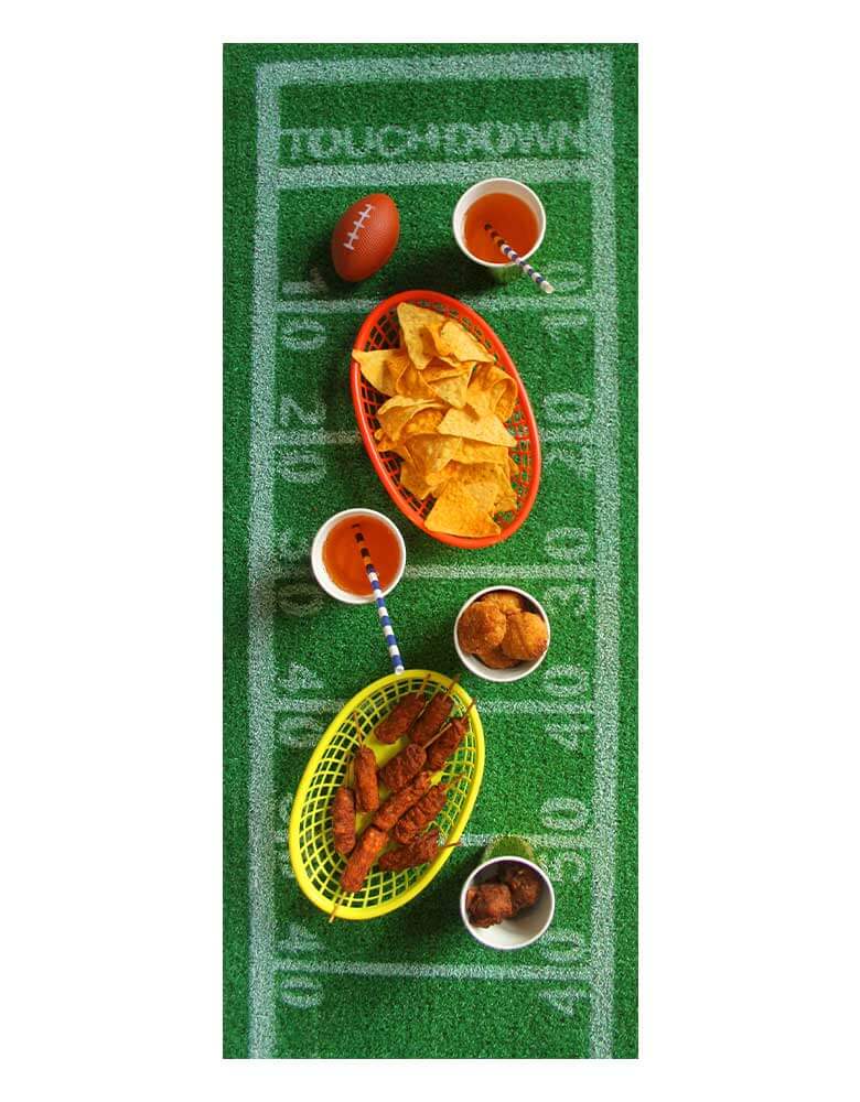 A overhead shot of a football party with Momo Party's 5ft x 16 inches Football Turf Table Runner by Talking Tables. On the table runner there are multiple football game snacks including chicken wings, chips and drinks. This grass runner features yard line markings and touchdown zones just like a football field. Perfect as a football party decoration for the Super Bowl party, game day or a kid's football-themed birthday party.