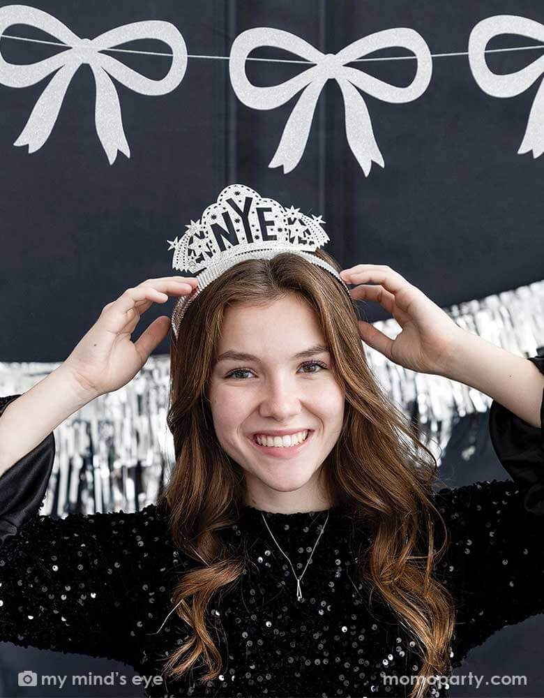 A young lady in a black sequin top wearing Momo Party's NYE silver glittered crown headband by My Mind's Eye. In her back hung a silver tinsel party banner and a silver glittered bow party garland, making this a modern and chic New Year's Eve countdown party decoration inspiration for this festive Holiday season.