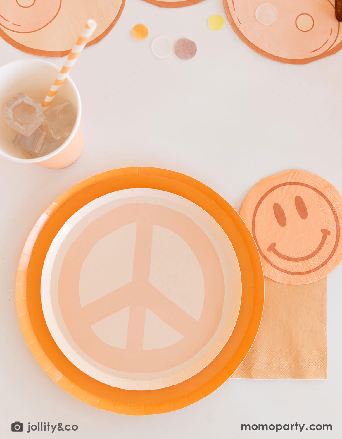 A neutral table setting with pastel colors of orange and peach peace sign shaped plate, a smiley face napkin, and a peach cup from Momo Party on a white table - a perfect inspo for a fun and groovy hippie love and peace themed party.