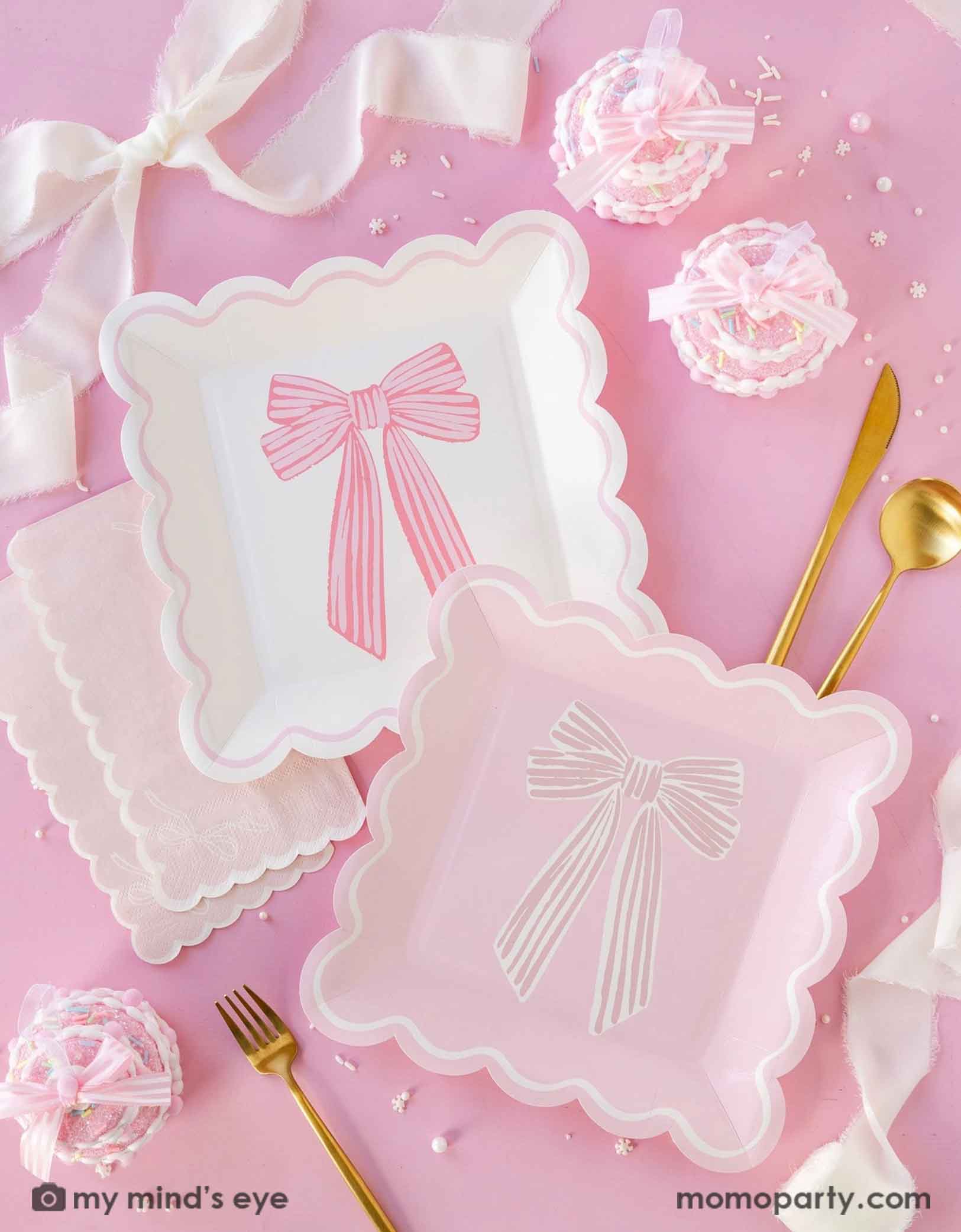 A pink tablescape featuring Momo Party's Momo Party's 9" pink and cream scalloped square plates with bow designs on by My Mind's Eye, along with scallop edged dinner napkins and gold metal utensils. With vintage pink cupcakes decorated with bows and some other decorated elements around the tablescape.