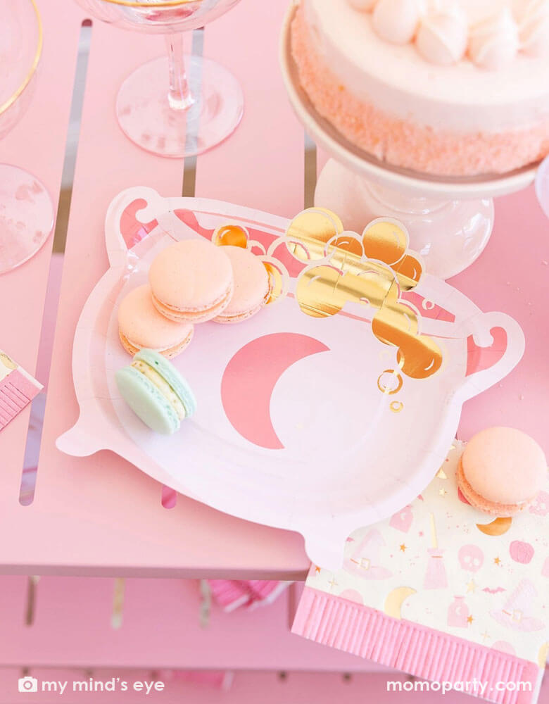 A pink Halloween party table featuring Momo Party's pink witch themed party supplies by My Mind's Eye including the pink cauldron shaped plate and the Hocus Pocus pink fringe napkin. On the table there are also some pastel colored macarons on the plate and the napkin which had a pink buttercream cake and some pink glasses next to them. This pink table is a perfect inspo for a fun and chic pink Halloween party for girls in the spooky season!