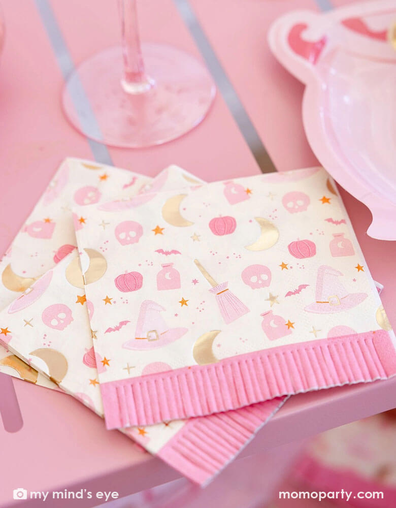 A close up shot of a pink table featuring Momo Party's pink witch Halloween party supplies including the pink Hocus Pocus fringe small napkins by My Mind's Eye. Featuring a fun "Hocus Pocus" pattern including witch themed designs like potion, witch's hats, skulls, bats, pumpkins, crescent moons, stars and fringe detailing, along with the pink cauldron shaped plates on the table, they're perfect for a chic pink witch themed Halloween celebration.
