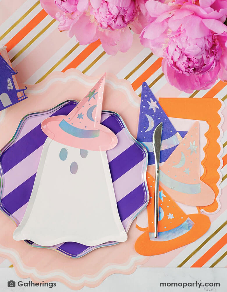 A festive pink Halloween party table featuring Momo Party's Bewitching ghost shaped plates, purple striped dinner plates, bewitching witch shaped hat napkins in three different colors of purple, orange and peach laid on a pink, orange and gold striped table runner. There are some beautiful peony flower arrangement in the middle of the table as the centerpiece, making this an adorable inspo for a chic and cute Halloween table for this spooky season!