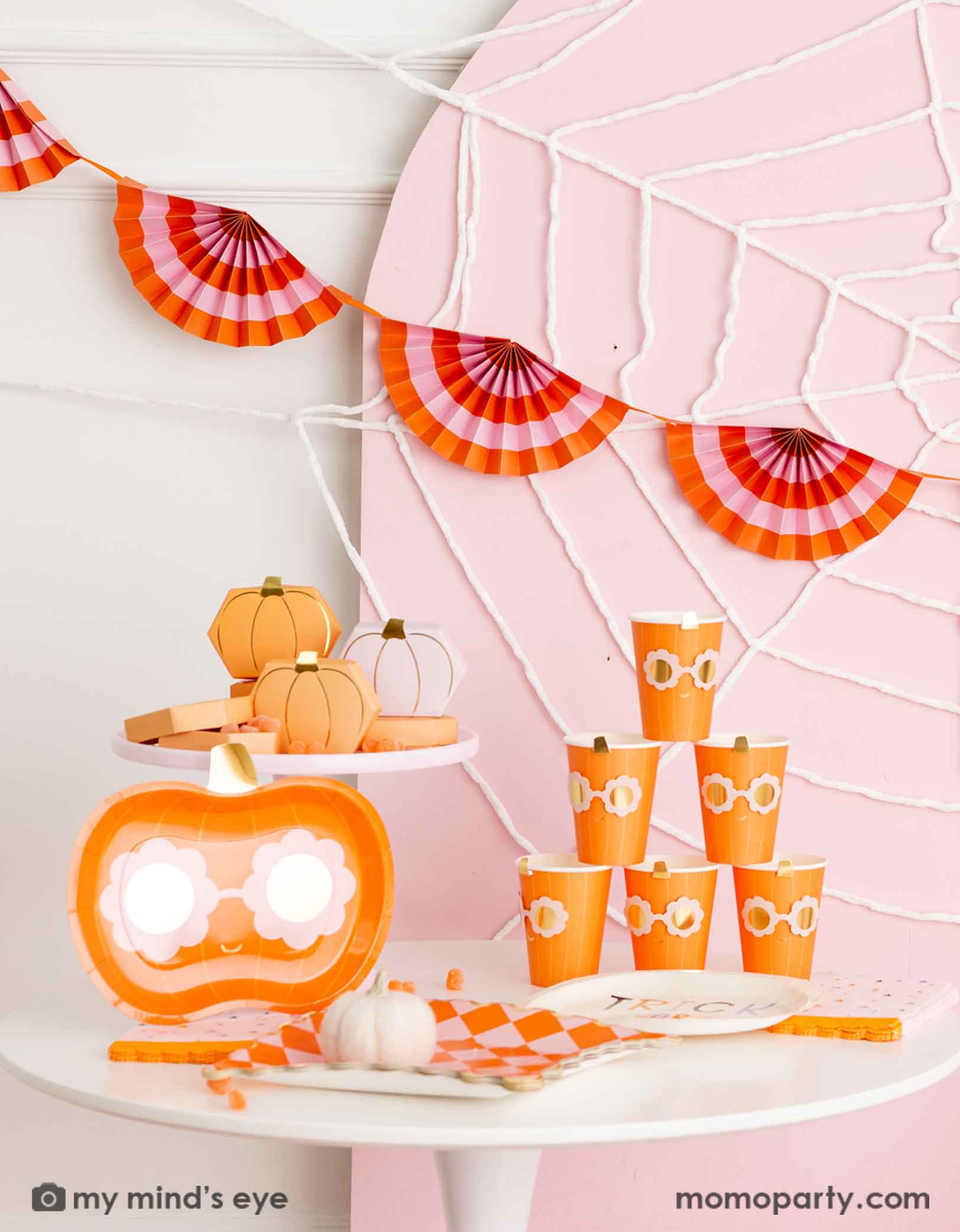 A Halloween-themed party setup with a table filled with Momo Party's groovy pumpkin-shaped plates with daisy-shaped sunglasses, groovy pumpkin party cups, orange and pink checkered plates and pumpkin shaped treat boxes on a pink cake stand. In the back there's a pale pink wooden curved backdrop board decorated with spider web. A sweet kid's friendly Halloween themed decoration.