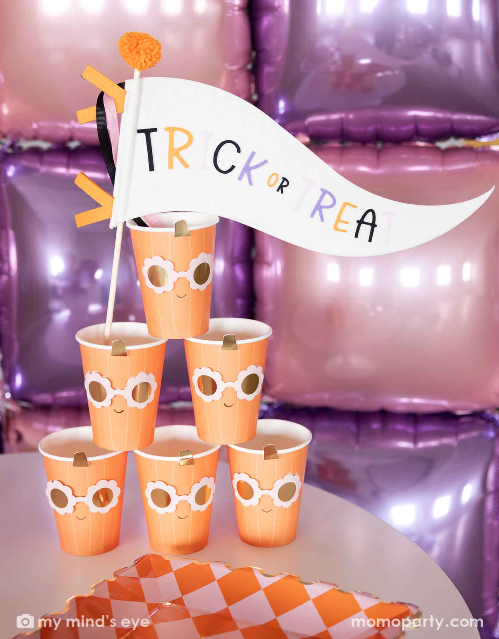 A stack of the orange pumpkin shaped paper party cups with daisy-shaped sunglasses from Momo Party with a 'Trick or Treat' felt party pennant on a Halloween inspired background with a foil balloon wall in pink and lilac. On the table there's a orange and pink checkered dinner plate.
