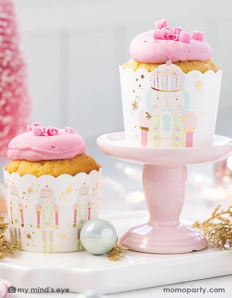 An adorable pink Christmas themed party table featuring pink cupcakes in Momo Party's gold Nutcracker food cups with gold foil accents on a small pink cupcake stand. Around the cupcakes there's some pink Christmas tree decorations in the back and some Christmas tree ornaments in gold and iridescent pastel colors. Making this a whimsical dreamy scene for a fabulous Holiday celebration.