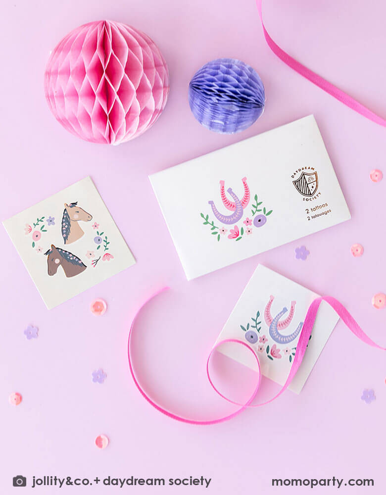 Momo Party's Pony Tales Temporary Tattoos by Daydream Society on a pink table with pink and purple mini honeycomb ball decorations around and flower shaped confetti. With some pink ribbons around the tattoo sheets, these temporary tattoos feature designs of ponies and horseshoes adorned with florals. They make perfect non-sugar party favors at your little girl's pony, cowgirl or rodeo themed party!
