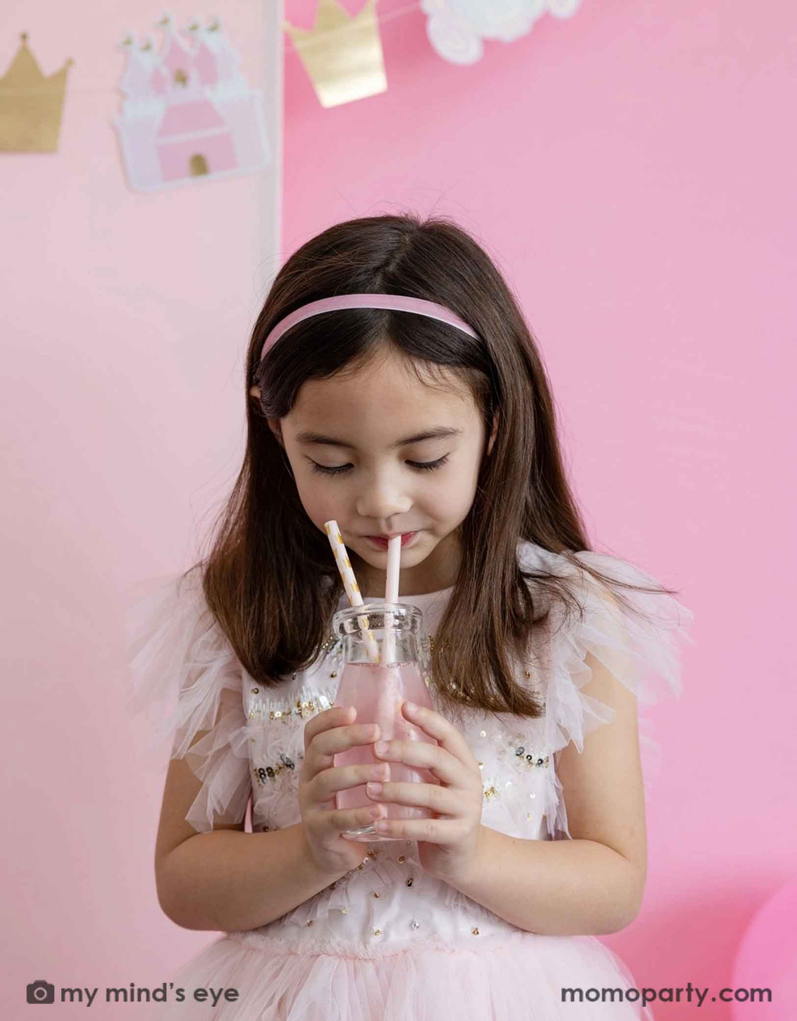 A young girl in a pale pink fairytale dress holding a pink drink with Momo Party's Princess Reusable Straws featuring gold crown pattern against a pink background, on the wall in the back there's also a princess themed party banner hung above along with some party balloons underneath.
