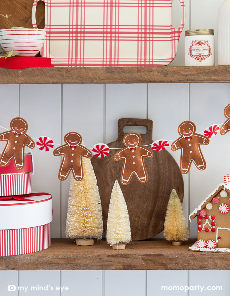 A kitchen self decorated with Momo Party's 7ft Puffy Felt Gingerbread and Candy Banner by My Mind's Eye along with some red striped bakeware including red gingham patterned bamboo serving tray, red striped mixing bowls and sugar can, along with a couple gift boxes with red and white striped wrapping and some white & beige colored bottle brush Christmas trees next to a gingerbread house. All makes it a great inspiration for Holiday home decor ideas.