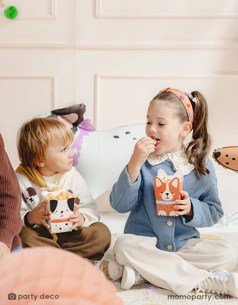 Two preschooler kids in a puppy dog themed birthday party sitting and holding Momo Party's puppy dog party snack boxes with adorable puppy design on them. They're eating the popcorn and chips from these cute puppy shaped treat boxes and having fun. In their back there are some cute dog shaped standing balloons.