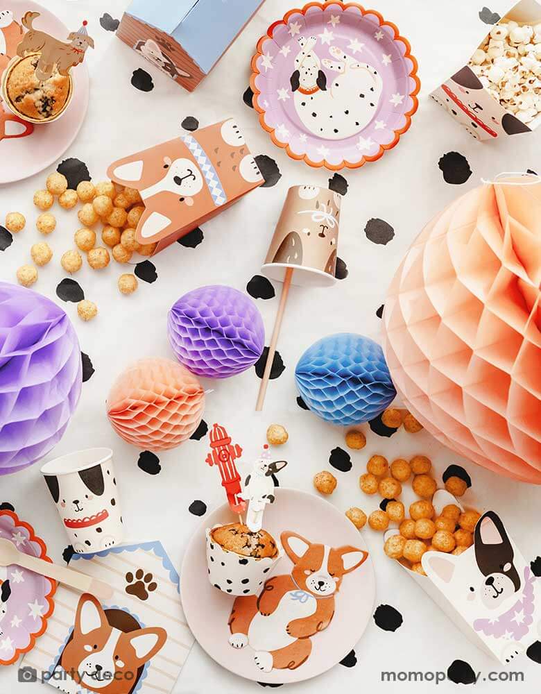 A festive puppy dog themed party tablescape featuring Dalmatian side plates, Corgi shaped napkins, dog shaped treat boxes, dog party cups, and cupcake toppers on a Dalmatian dog printed tablecloth. Around the plate there are some honeycomb ball decorations in soft pastel colors, making this an adorable inspiration for a cute dog puppy themed celebration.