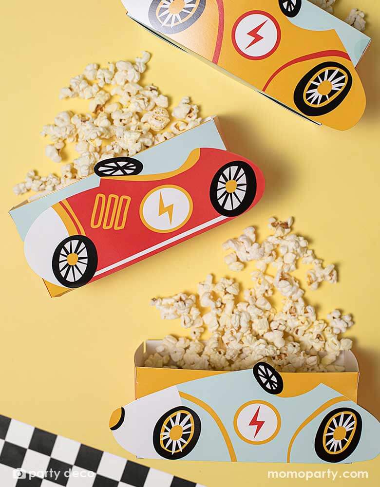 A festive bright yellow party table features three of Momo Party's race car snack boxes by Party Deco. These race car treat boxes are filled with popcorn, next to a race car flag checkered table runner, perfect for kid's car themed celebrations.