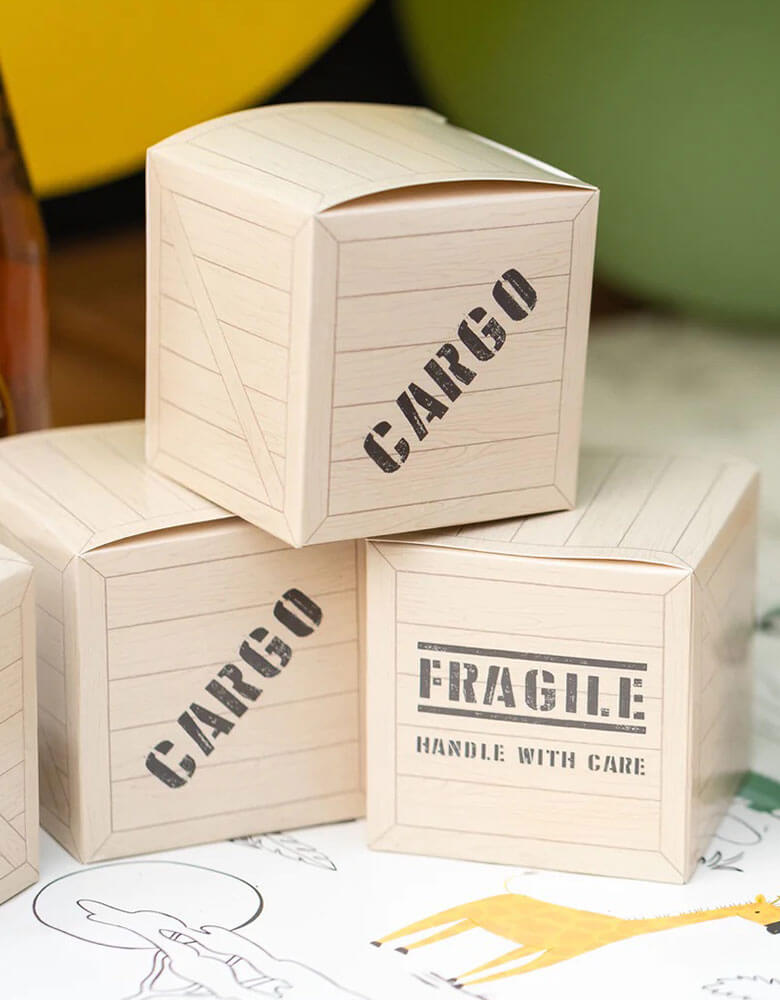 A safari themed party table featuring 3 of Momo Party presents 3" x 3' safari adventure cargo favor boxes by My Mind's Eye. Designed to resemble cargo boxes, each favor box has "Cargo" & "Fragile, handle with care" on the sides. These favor boxes are the perfect way to add some adventure to your safari themed birthday celebrations!