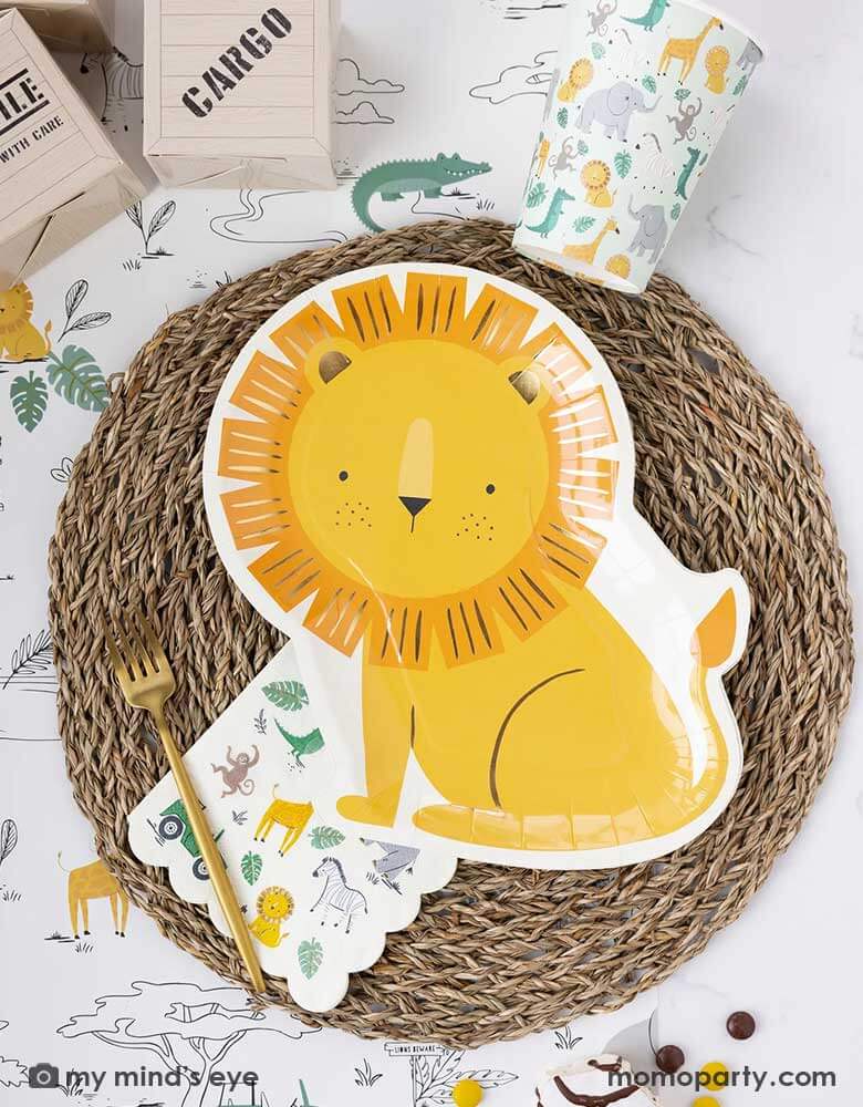 A safari adventure themed party table featuring Momo Party's 9" x 11" lion shaped plate laid on top of a round bamboo weaved round tablemat which is placed on My Mind's Eye's safari adventure paper table runner featuring safari animals and safari landscape. Around the plate are a mint party cups with lots of safari animals and the safari animal 5" x 5" small napkin. With My Mind's Eye's cargo favor boxes and zebra cakes on the table, makes this tablescape a great inspiration for kid's safari themed party.
